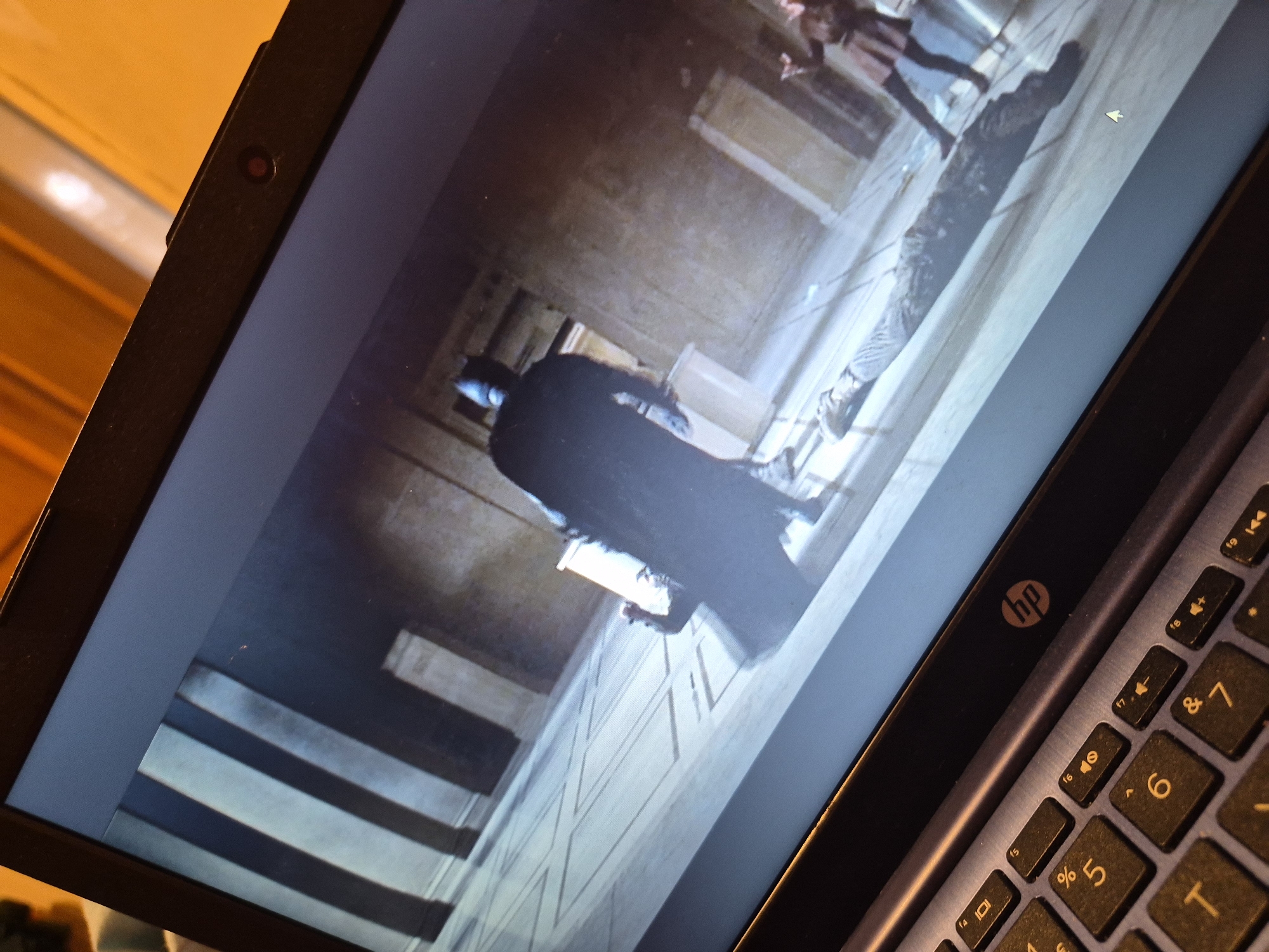 The image shows a laptop screen displaying a scene from what appears to be a movie or television show, with Batman in the foreground. The scene takes place indoors, in a large, open room with tall columns and a patterned floor. A person is lying on the floor, seemingly unconscious, while Batman stands over them. Another person is visible in the background to the right. The laptop itself is positioned on a surface, and the keyboard is partially visible. The image is taken from a high angle, looking down at the laptop. There is no information available in the image that would allow for the identification of a location in the city of Sandwell, United Kingdom.