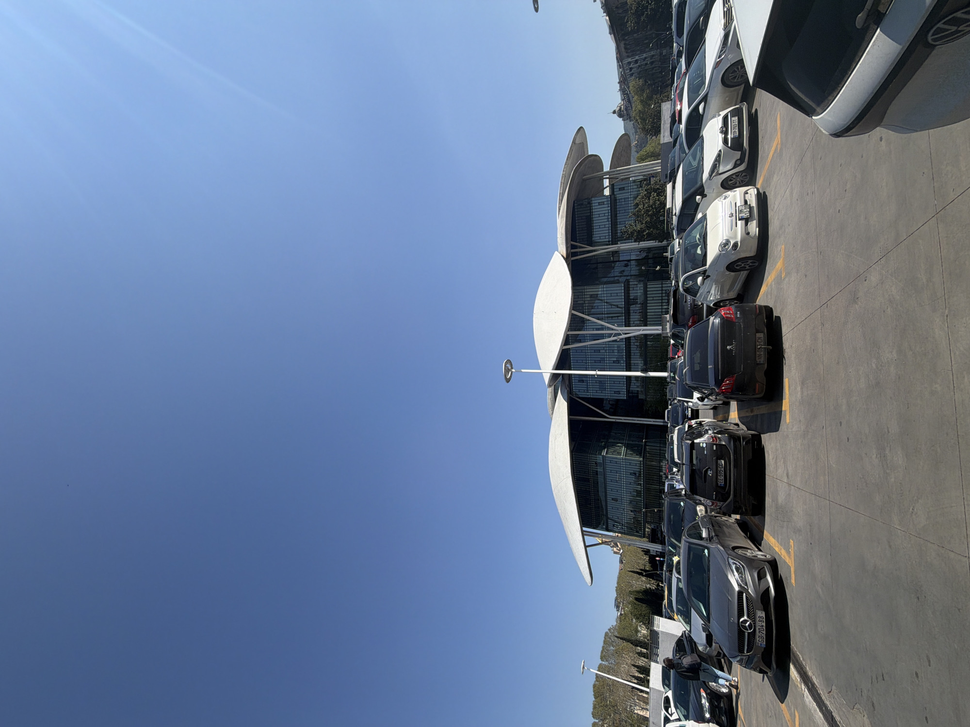 The image, viewed in its correct orientation, captures a bright, sunny outdoor scene of a large parking lot in front of a distinctive modern building. The building, identifiable as the Public Service Hall in Tbilisi, Georgia, features a multi-story glass facade topped with several white, curved, shell-like roof structures.
The parking lot is filled with numerous parked cars of various makes and models, including a white Fiat 500, a white Toyota Yaris, and a grey Mercedes-Benz SLK, all neatly aligned within yellow-marked parking spaces. Sunlight is strong, casting distinct shadows from the vehicles and the building, suggesting midday. The sky is clear and blue, devoid of clouds.
One person, wearing a blue jacket and jeans, is partially visible near a parked car on the left side of the frame. In the distant background, beyond the building, are hillsides dotted with trees and other structures. The scene depicts a static moment at a busy public or commercial facility.
Visible text from license plates includes "GB-704-BB," "EE-595-CC," "NN-053-CN," and "TP-783-TT."