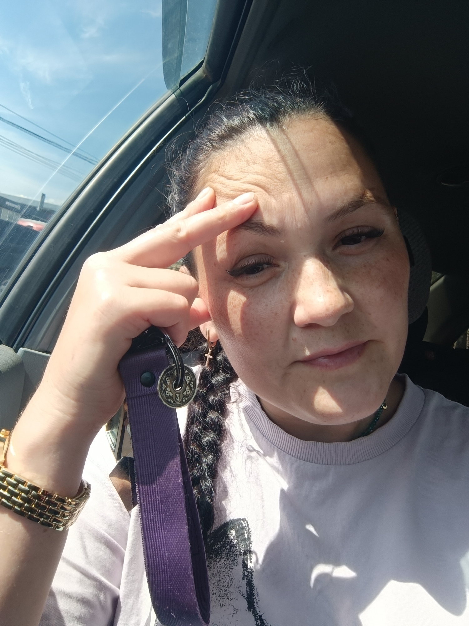 The image shows a woman in the front passenger seat of a car. She is looking at the camera and has her left hand on her forehead, with her index finger pointing to her temple. She is wearing a light purple t-shirt and a gold watch on her left wrist. A purple lanyard with a keyring is also visible. The background shows the sky and part of the car's interior. The scene appears to be a selfie taken during the day, possibly while traveling. There is no visible indication of the specific location within the city of Chaikovsky, Russia.