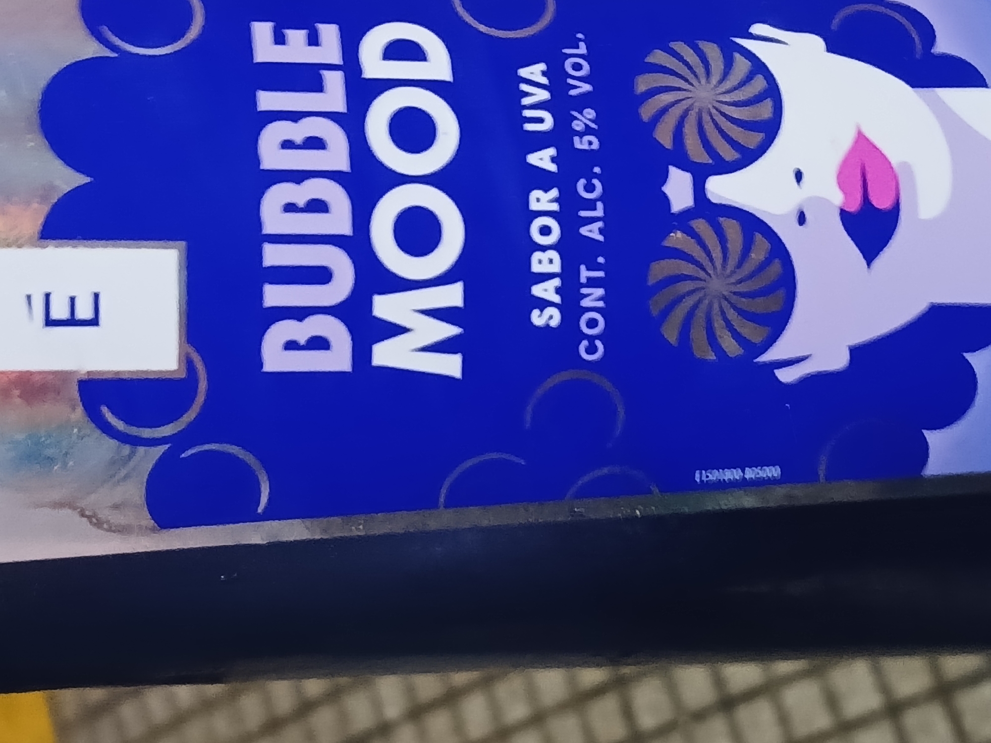 A close-up image displays a portion of a product's packaging, likely a beverage. The packaging is predominantly blue with a bubbly design. The words "BUBBLE MOOD" are printed vertically in large, white, sans-serif font. To the right of the text, there's a cartoonish white figure with stylized eyes resembling pinwheels or swirls. The figure also features a pink tongue sticking out. Text below "BUBBLE MOOD" reads "SABOR A UVA CONT. ALC. 5% VOL." in a vertical orientation. A small white label on the left edge shows the letter "E" in a sans-serif font. A code "1504000005000" is printed in a small font towards the bottom right. The image seems to be taken indoors, with part of a dark surface visible at the bottom. The context or location within Buenos Aires, Argentina, is not visually discernible from the image provided.