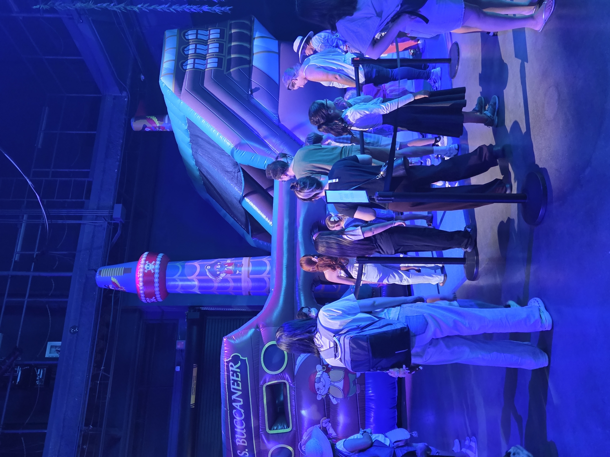 The image depicts a group of people queuing in front of an inflatable pirate ship attraction, likely at an indoor amusement park or event space. The attraction is brightly colored with shades of purple, blue, and pink, featuring pirate imagery. A prominent inflatable tower on the left side of the ship is decorated with a pirate character and a skull and crossbones. To the right, a large inflatable slide rises from the ship. The area is dimly lit with a strong blue hue, suggesting artificial lighting. People of various ages are visible, dressed in casual clothing. Some individuals are wearing masks. The ground appears to be a smooth, dark surface reflecting the ambient blue light. Stanchions with black ropes are set up to guide the queue. The overall scene conveys a sense of anticipation and entertainment.