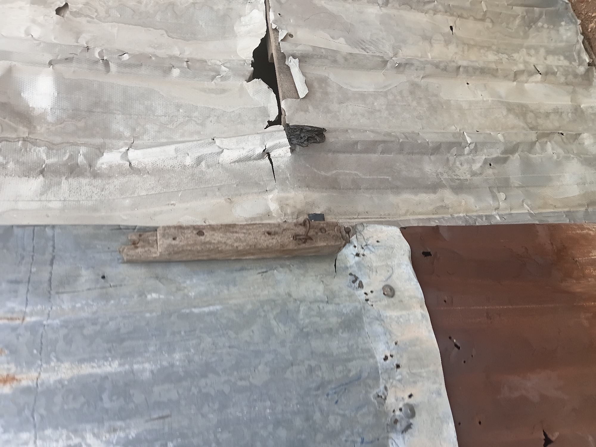 The image displays a close-up view of a damaged corrugated metal structure, likely a wall or roof, in Jalingo, Nigeria. The primary subjects are sheets of corrugated metal, some appearing weathered and rusted, others covered in a white material that is peeling and torn in places. There is a visible wooden beam affixed to the structure, with metal fasteners. The scene suggests disrepair and neglect, with significant damage to the metal sheeting, including holes and cracks. The lighting appears natural, possibly indicating daytime, but the weather conditions are not discernible. The overall impression is one of decay and the passage of time, with no people or specific activities depicted. The texture of the materials is rough and uneven, emphasizing the dilapidated state of the structure. The color palette is muted, dominated by grays, whites, and rust tones, with a darker reddish-brown section of metal visible on the right. No text is visible in the image.