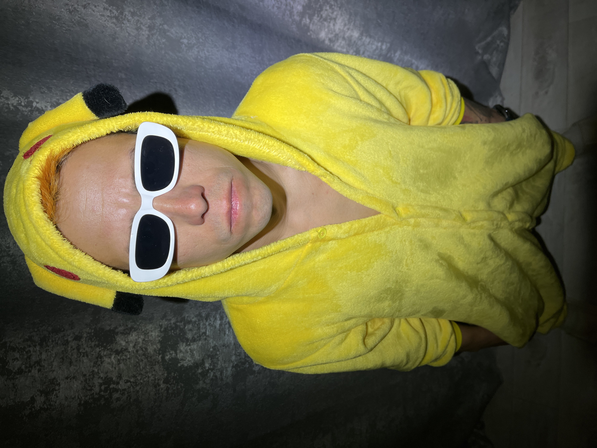 A person wearing a yellow Pikachu onesie and large white sunglasses is lying on a textured gray surface. The person's head is positioned toward the top left of the frame, and their body extends diagonally toward the bottom right. The background appears to be a gray curtain or fabric. The photograph was taken indoors under mixed lighting conditions, creating shadows on the right side of the frame. There is no information in the image to suggest that the location is Chaikovsky, Russia.