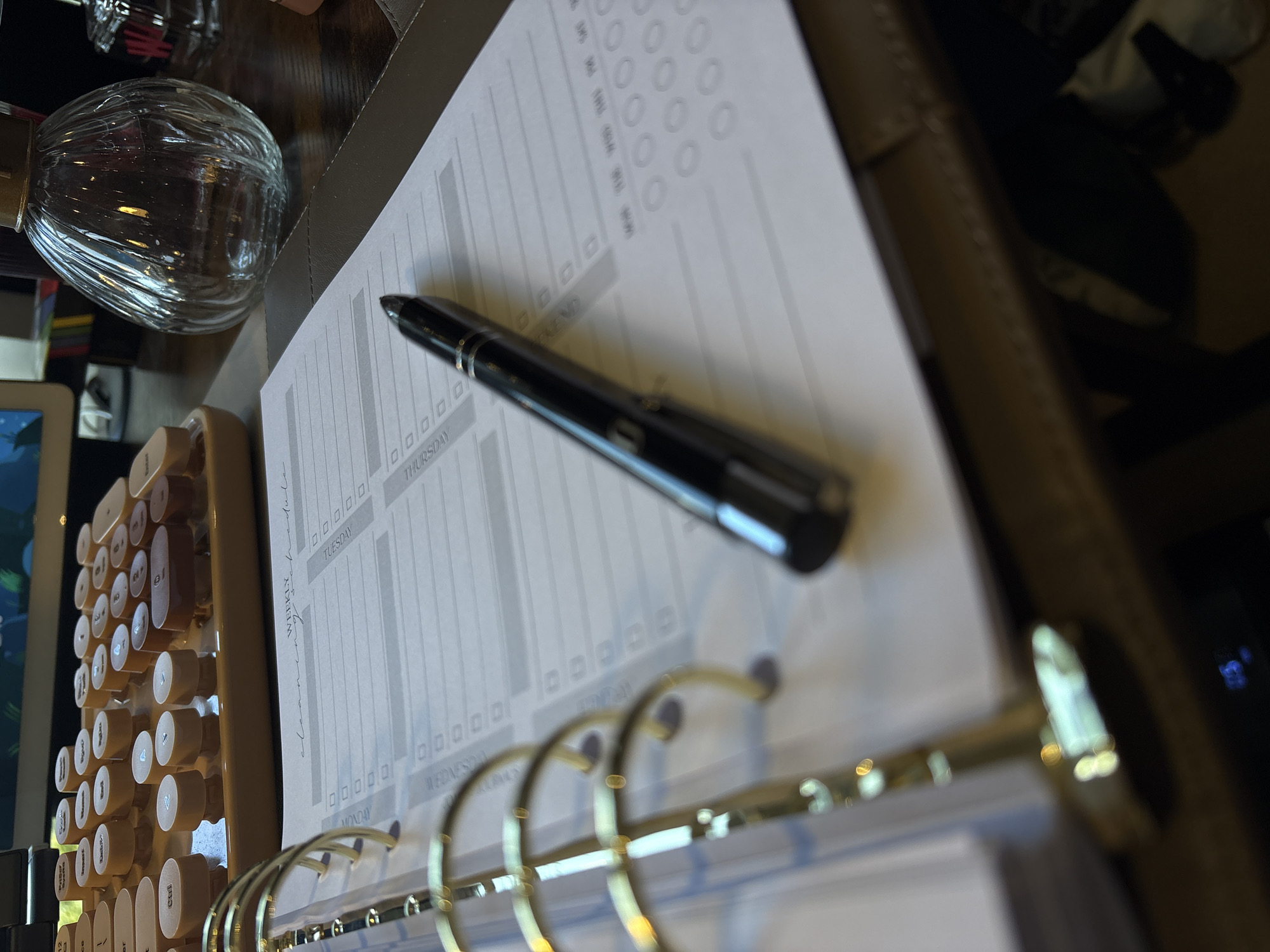 The image shows a close-up of a desk with various objects. A planner with gold rings lies open, displaying a page titled "WEEKLY cleaning schedule." The days of the week, Monday through Thursday, are printed on the page. A black pen rests on the page. To the left of the planner is a cream-colored mechanical keyboard with round keys, and behind that is a partially visible screen. A glass bottle-shaped object is also present, and a glimpse of a person or chair is visible in the upper right corner. The location is not identifiable as being in Ipswich, United Kingdom.