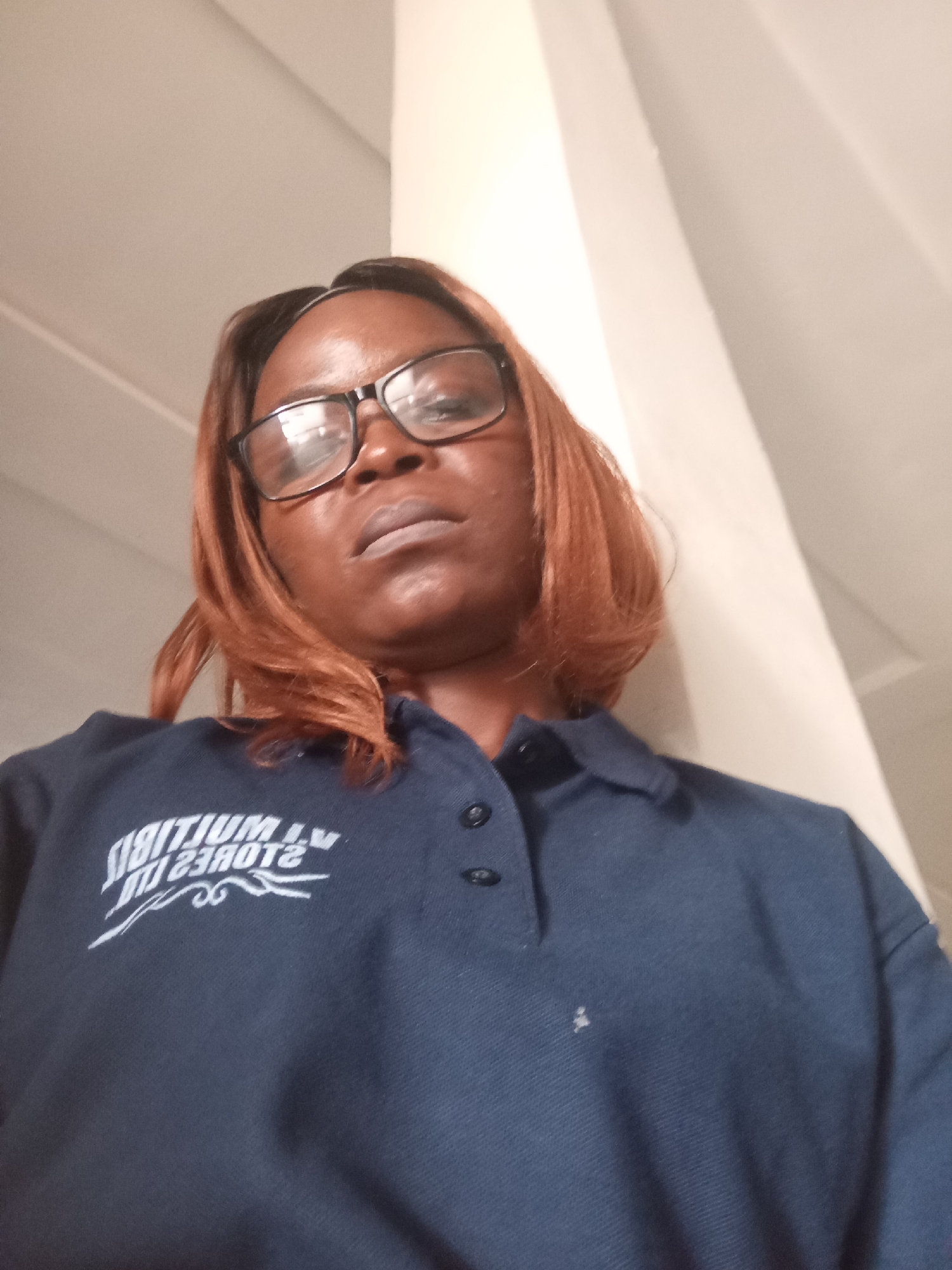A low-angle, medium shot captures a Black woman from the chest up, looking slightly upwards and towards the viewer. She is wearing a dark blue polo shirt with a white logo on the left side of the chest. The logo features text that appears to be in a stylized font, with some elements mirrored or reversed. The woman has medium-length, auburn-colored hair that frames her face. She is wearing black-rimmed glasses. Her skin tone is dark, and her lips are slightly parted. The background consists of a wall that appears to be painted a light, neutral color, with a distinct shadow cast from a vertical object, possibly a pillar or corner of a building. The lighting suggests an indoor environment, possibly under artificial light, as there are reflections on her glasses and the shirt. The image quality is moderate, with some graininess. The specific location of Mayo Dasa, Nigeria, is not visually indicated within the image itself.