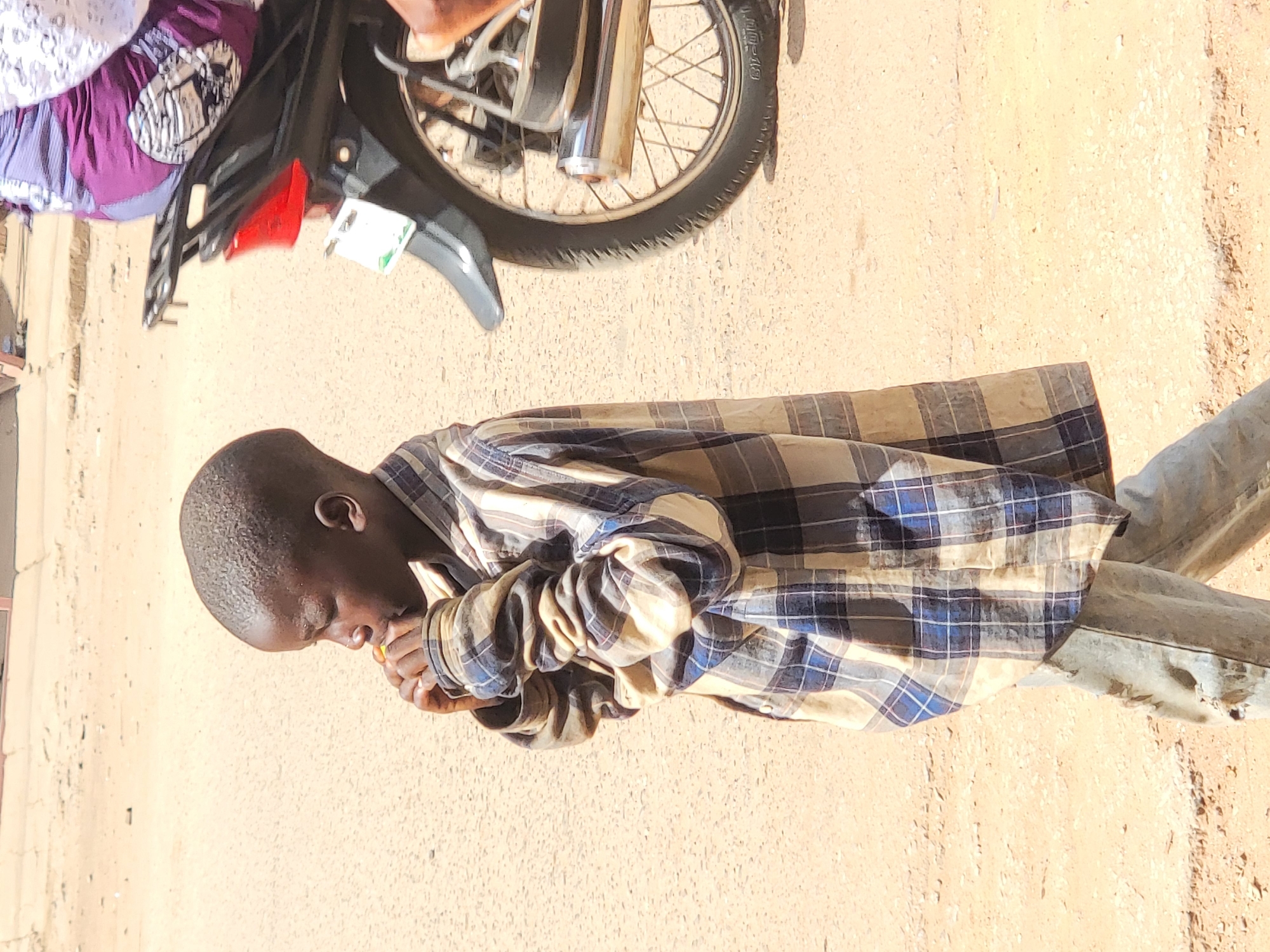 A young man with dark skin is standing on a dirt road in Maiburiji, Nigeria. He is wearing a blue and tan plaid shirt and faded gray pants. He is looking down and appears to be eating something he is holding in his hands. To his left, partially visible, is a parked motorcycle with a passenger wearing a purple and white patterned garment. The ground is a light brown color, and the scene is bathed in sunlight.