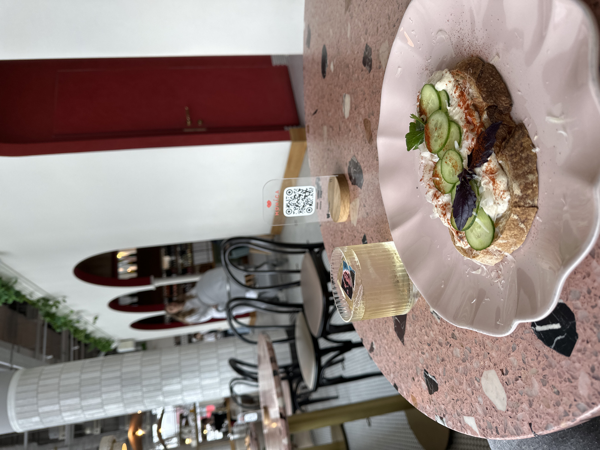 Here's a detailed description of the image:
**Setting:** The photo was taken at a cafe. The location is likely in Nizhny Novgorod, Russia based on the 'NOmica' branding on the QR code stand.
**Objects:** The primary objects include:
* A light pink plate with a culinary dish on it. The dish appears to consist of a slice of bread topped with a creamy spread, cucumber slices, herbs (parsley and basil), and seasoning (likely paprika).
* A glass of clear liquid (likely a beverage).
* A small, transparent QR code stand with a wooden base. The stand has "NOmica" written on it.
* A decorative terrazzo-style table with a pink and black stone pattern.
* Several black chairs are visible in the background.
**Environment:**
* The cafe has a modern interior design. A white, tiled wall is visible, along with red accents (doors and ceiling).
* Some greenery hangs from the ceiling.
**People:**
* There is a blurry figure of a person in a gray shirt seated in the background, near the chairs.
**Actions/Interactions:** The image is a still life, likely staged to showcase the culinary offering of the cafe. No direct actions or interactions are depicted, but the presence of a QR code suggests a promotional context (i.e., to access a menu or online information).