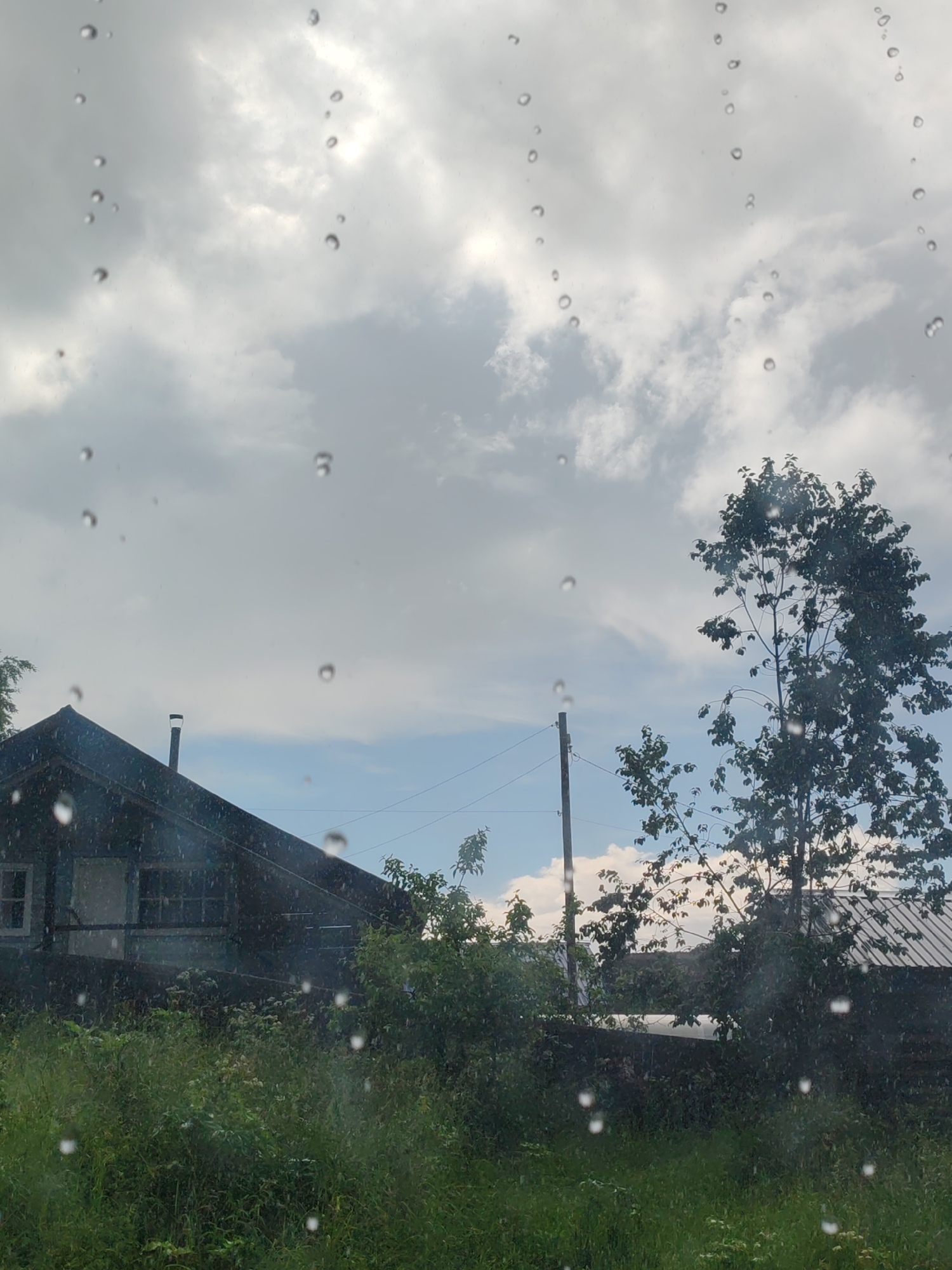 This is an outdoor scene, likely captured from inside a vehicle or building due to the presence of raindrops on the foreground glass. The sky is overcast with dramatic, grey clouds interspersed with patches of blue sky. A dark, wooden house with a gabled roof and a chimney is visible on the left side of the image. In front of the house, dense green foliage and tall grass cover the ground. To the right, a large, leafy tree stands tall against the sky, and another smaller tree is visible behind it. A utility pole with power lines crosses the scene horizontally in the background. The overall impression is of a rural or semi-rural setting, possibly in the city of Chukhlom, Russia, on a cloudy, possibly rainy day.