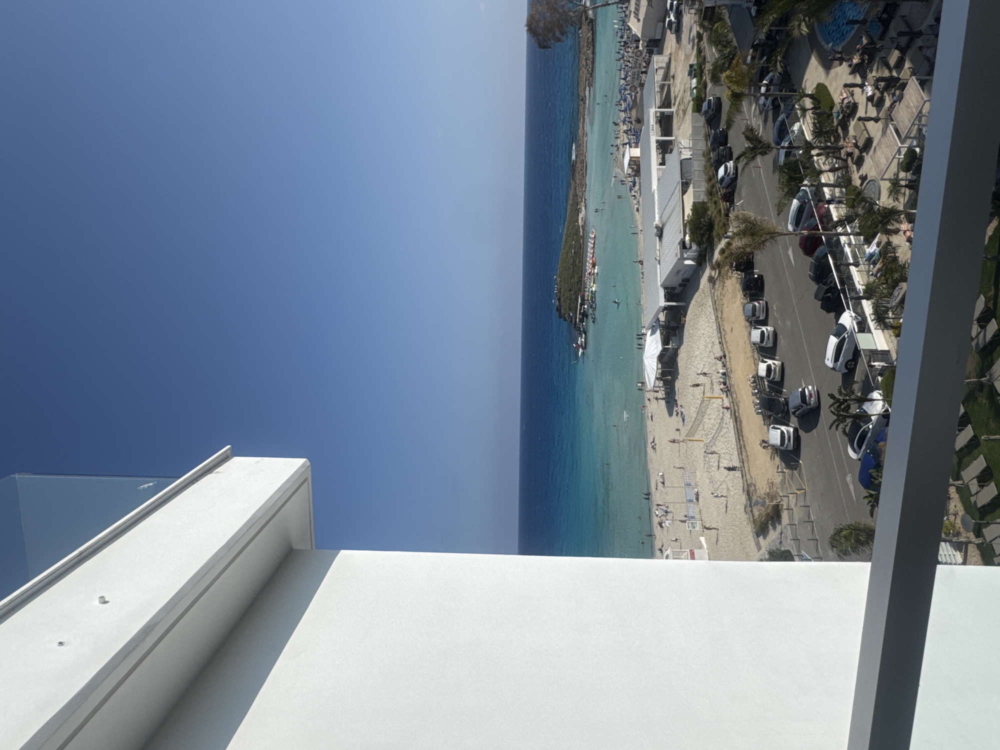 An aerial view, taken from a high-rise balcony, captures a vibrant coastal scene under a clear blue sky. The foreground features a stark white balcony wall with a glass railing, providing a high vantage point over the resort below.
The left side of the image is dominated by an expanse of clear blue sky, transitioning subtly in shade from left to right. The right half of the image showcases a bustling seaside area. A broad stretch of turquoise sea meets a sandy beach, with the water deepening to a darker blue further from the shore. Numerous small boats are visible both docked and scattered across the water.
The sandy beach is dotted with many small figures of people engaging in various leisure activities, such as swimming, sunbathing on sun loungers, and walking. Beach umbrellas and a volleyball net are also present. Adjacent to the beach, a paved road runs parallel to the coastline, lined with parked cars and interspersed with green palm trees. Beyond the road, several white-colored buildings, possibly hotels or residential units, rise up, some featuring multiple stories. A visible pool area with more sun loungers and umbrellas is situated among these buildings.
The bright, strong sunlight indicates a clear mid-day, casting defined shadows across the landscape. The overall atmosphere is one of a lively, sunny vacation destination. Visible text includes "NISI BAY" on a blue banner near the beach and "LUNA" on a building sign.