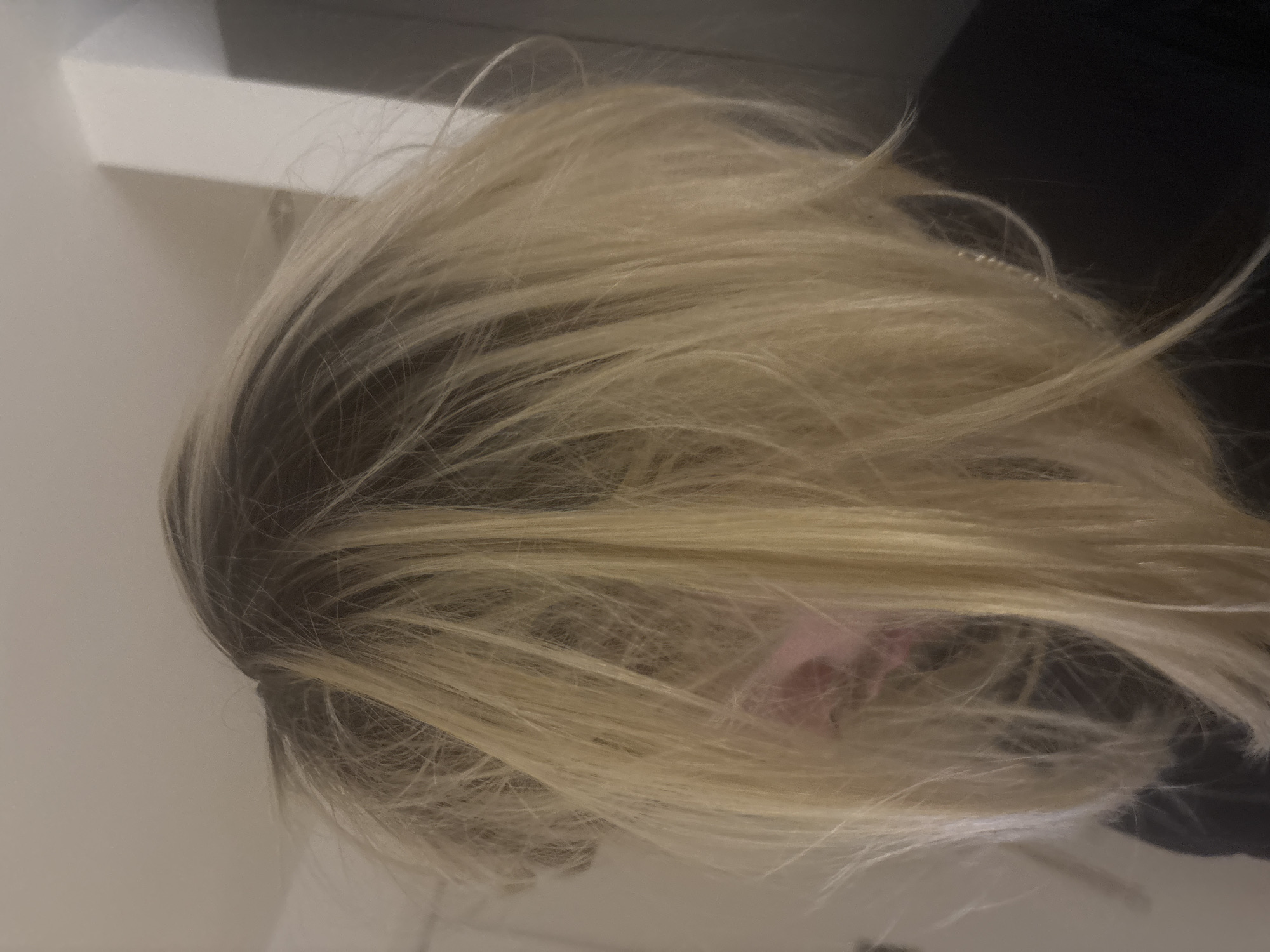 The image shows a close-up of a person's head with long, blonde hair that is parted on the side, revealing darker roots. The hair appears to be slightly disheveled. A portion of the person's face, including the nose and cheek, is partially visible through the hair. The background is indistinct but suggests the presence of a wall on the left and a dark object on the right, possibly clothing. There is no information indicating the location is in London, United Kingdom.