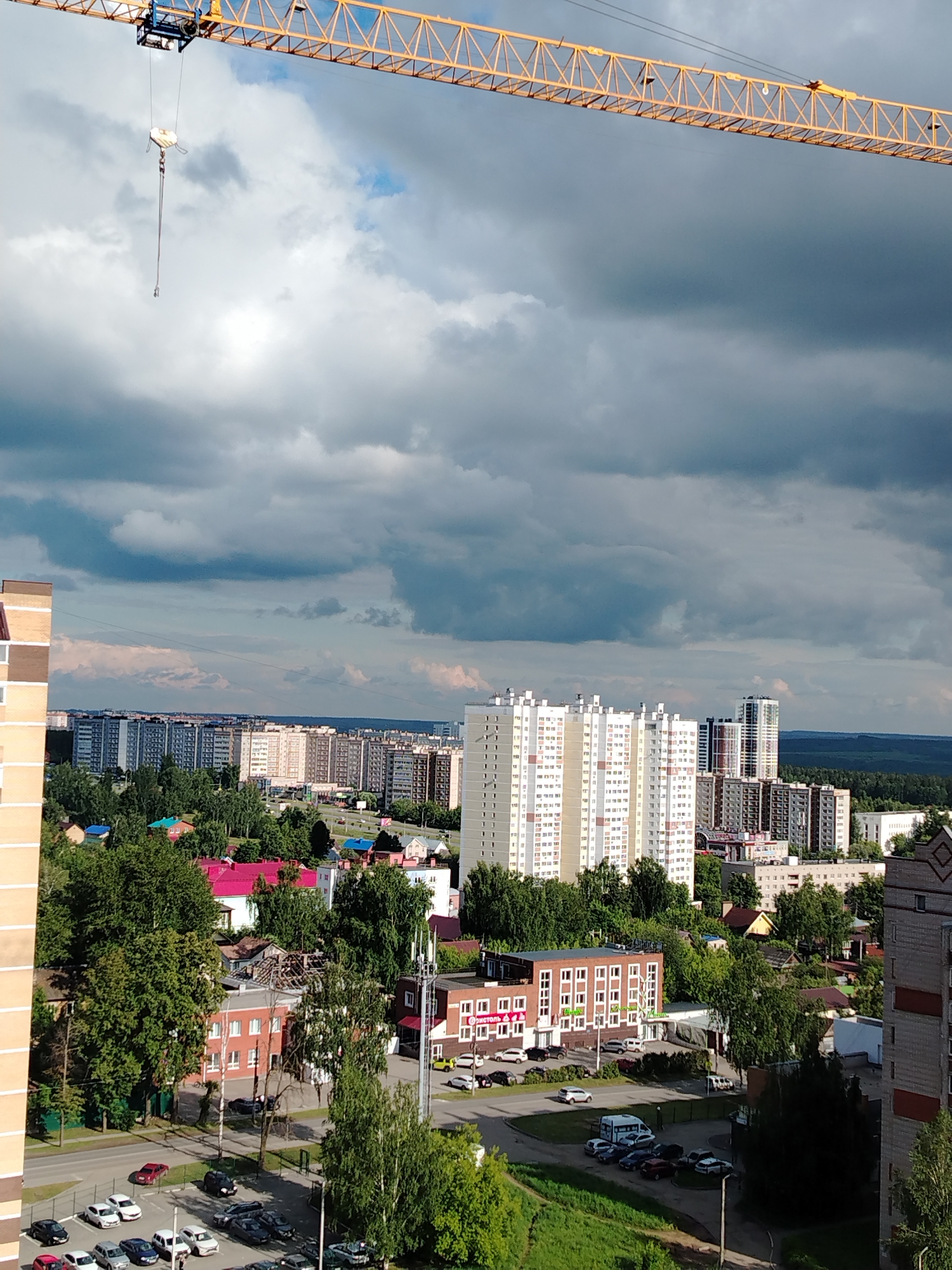 An elevated, wide-angle view captures a sprawling urban landscape under a heavily clouded sky. The scene is set in Izhevsk, Russia, showcasing a mix of residential and commercial structures amidst abundant greenery.
Dominating the top of the frame is the prominent yellow boom of a tower crane, extending diagonally from the upper left. Its hoist block and hook hang down towards the center-left, suggesting ongoing construction activity, although no workers are visible.
The sky is filled with large, dark grey clouds, indicating overcast weather. Patches of lighter cloud and hints of blue sky are visible, suggesting the sun might occasionally break through. The overall lighting is diffuse, consistent with a cloudy daytime setting.
Below the crane, the city unfolds. Numerous multi-story apartment buildings, varying in architectural style and age, populate the midground and background. Some are modern high-rises with light-colored facades (white, beige), while others appear to be older, more uniform blocks. Interspersed throughout the urban fabric are dense clusters of green trees, lining streets and surrounding buildings, creating a verdant feel.
Roads wind through the city, with several cars parked in lots and traveling on streets. A distinct white van is visible in one parking area. In the midground, a red brick building stands out, featuring multiple windows and a sign with the Russian text "ДИОНСТОЛЬ" (Dionstol).
In the far background, a dark green forested area or range of hills stretches across the horizon, indicating the city's proximity to natural landscapes. The overall impression is of a lively, developed city, adapting to its environment under a dramatic sky.