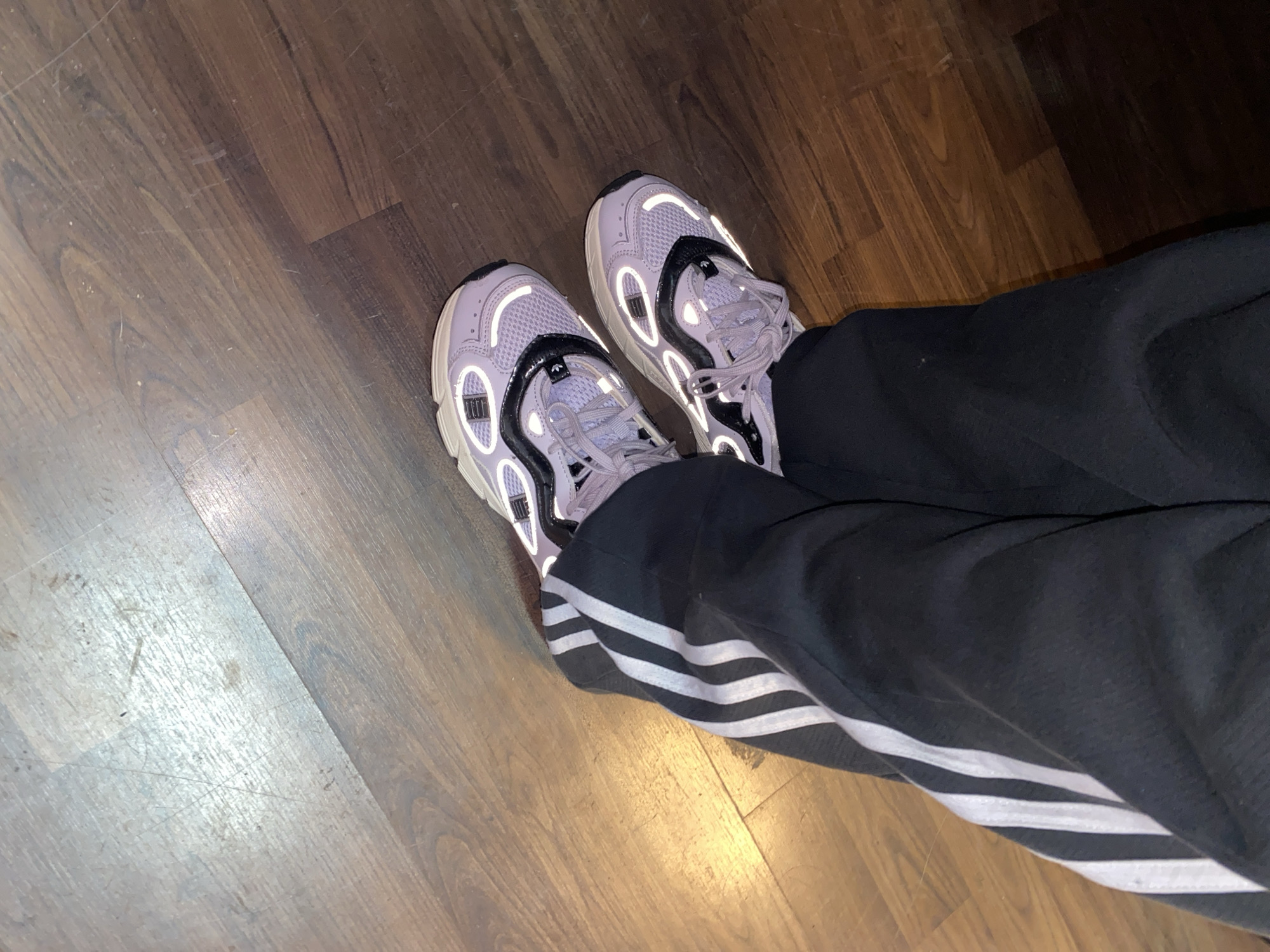 The image shows a person wearing gray athletic shoes with black and white detailing and black Adidas sweatpants with white stripes, standing on a hardwood floor. The location is not discernible from the image. No actions or interactions are visible.