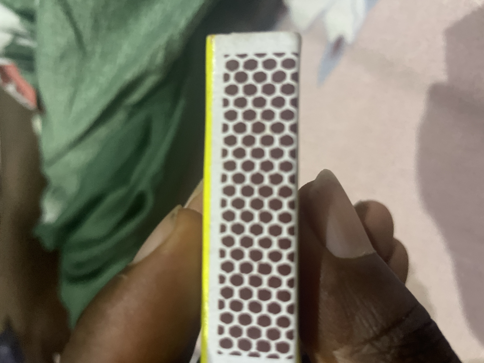 A close-up, top-down shot shows the side of a matchbox being held by a person's fingers. The matchbox has a yellow border and a white side with a brown, hexagonal, scratch-resistant surface. The background is blurred, showing green fabric and a pinkish surface. The image is taken in Jalingo, Nigeria.
