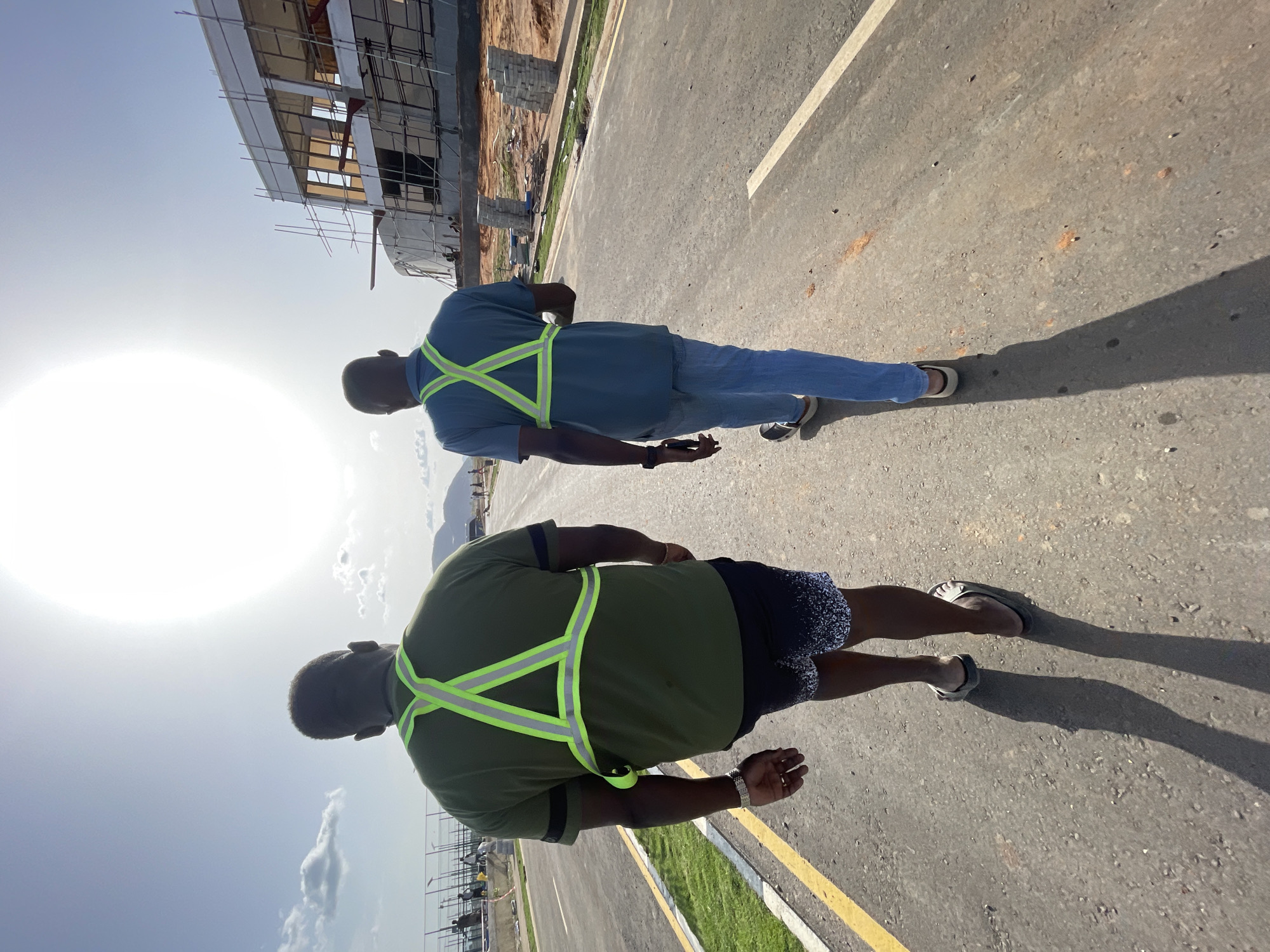 Two men wearing safety vests are walking on a paved road. The road appears to be newly constructed, as there is adjacent construction visible in the background. The photograph was taken during the day in bright sunlight. The location is identified as Angwan Sayawa, Nigeria.
