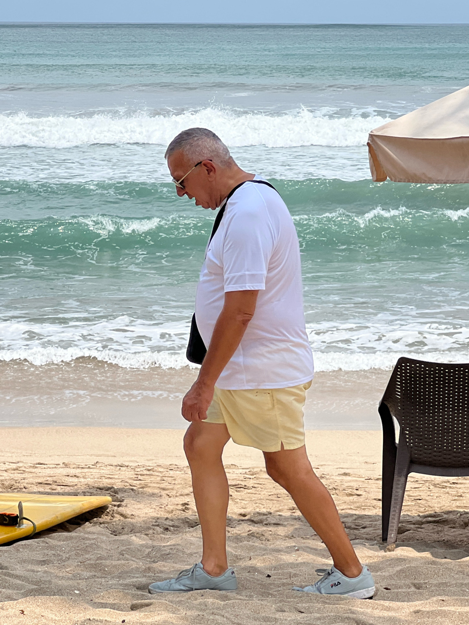 An elderly Caucasian man wearing a white t-shirt, light yellow shorts, and gray sneakers is walking on a sandy beach in Kuta, Indonesia. He has gray hair and is wearing sunglasses. A black bag is slung over his left shoulder. The ocean with moderate waves is behind him. To his left, a yellow surfboard is partially visible on the sand. To his right, a dark brown plastic beach chair and a portion of a beige umbrella are present. The sky is overcast and the ocean water is a greenish-blue color.