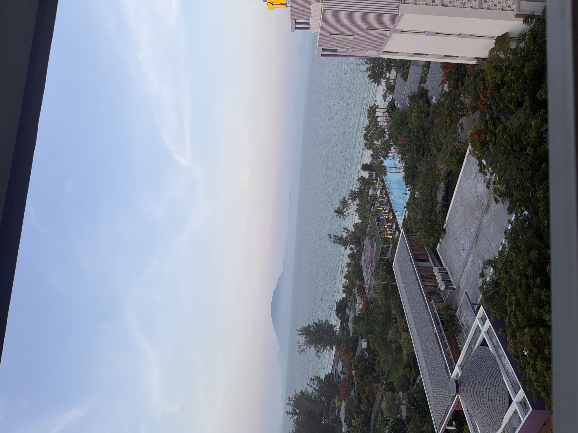 The image presents a high-angle, panoramic view from an elevated position, likely a balcony or window, overlooking a coastal resort in Dong Ninh Hoa Ward, Vietnam.
The upper portion of the frame features a pale blue sky with faint, wispy clouds. Below the sky, a wide expanse of calm, light blue-green ocean dominates the mid-ground. Along the distant horizon, several layers of hazy, bluish-grey mountains or landmasses are visible, with a prominent, larger mountain centrally positioned. A small boat is discernible on the water in the mid-distance.
The coastline reveals a sandy beach populated by numerous small figures, indicating human presence. Adjacent to the beach and extending into the foreground, there is dense, lush green vegetation, including many palm trees and other tropical trees, some displaying bright red or orange blossoms.
Within the resort complex, a large, blue-tiled swimming pool is visible in the mid-foreground, containing several individuals. The pool area is equipped with lounge chairs and umbrellas. Various buildings with distinctive architecture are scattered throughout the resort. On the far right, a multi-story building with a white facade and purple vertical stripe accents is partially visible, featuring a yellow sign with a partial "T" on its roofline. In the lower-left foreground, a building with a unique grey, conical shingled roof and purple-trimmed concrete base is present. Other low-rise buildings with grey shingled roofs and purple details are interspersed among the trees. Air conditioning units are visible on the flat roof of one building in the mid-foreground. The scene is illuminated by soft, diffused daylight.