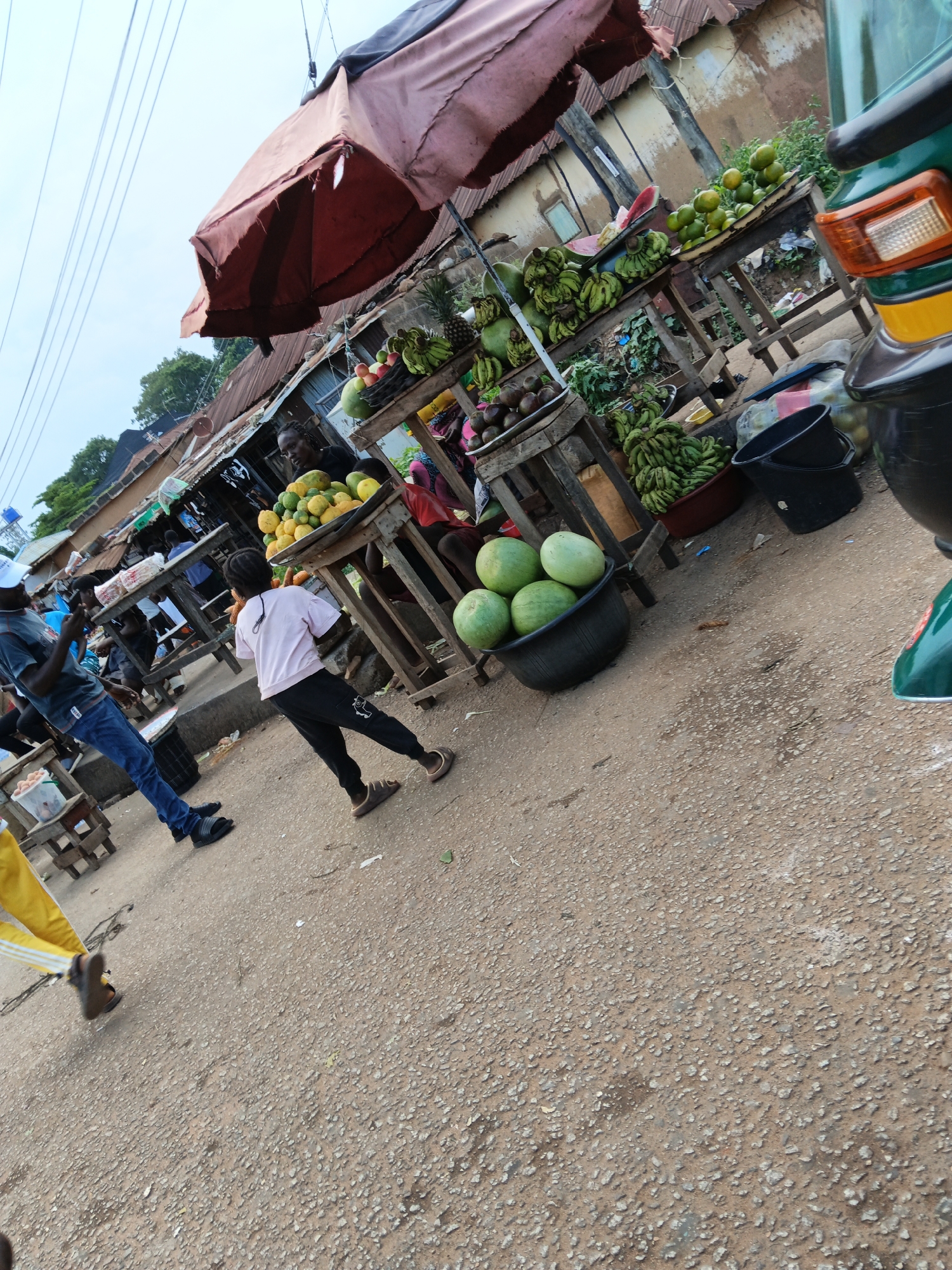 The image depicts a street market scene in Bukuru, Nigeria. A collection of fruit and vegetable stalls line a dirt road, with various produce displayed on wooden tables and in crates. Prominently featured are large bunches of green bananas, piles of yellow and orange fruits resembling mangoes or papayas, and several watermelons in a black tub. A young girl in a pink shirt and black pants is walking across the scene. To the left, a man in a blue shirt and jeans stands with his phone. In the background, there are several people at the market stalls, and rudimentary structures with corrugated metal roofs. An overhead canopy, possibly an umbrella, is partially visible. A green and yellow motorized tricycle is parked to the right, partially obscuring some of the market stalls. The sky is overcast, and power lines are visible overhead. The ground is a mixture of dirt and gravel.