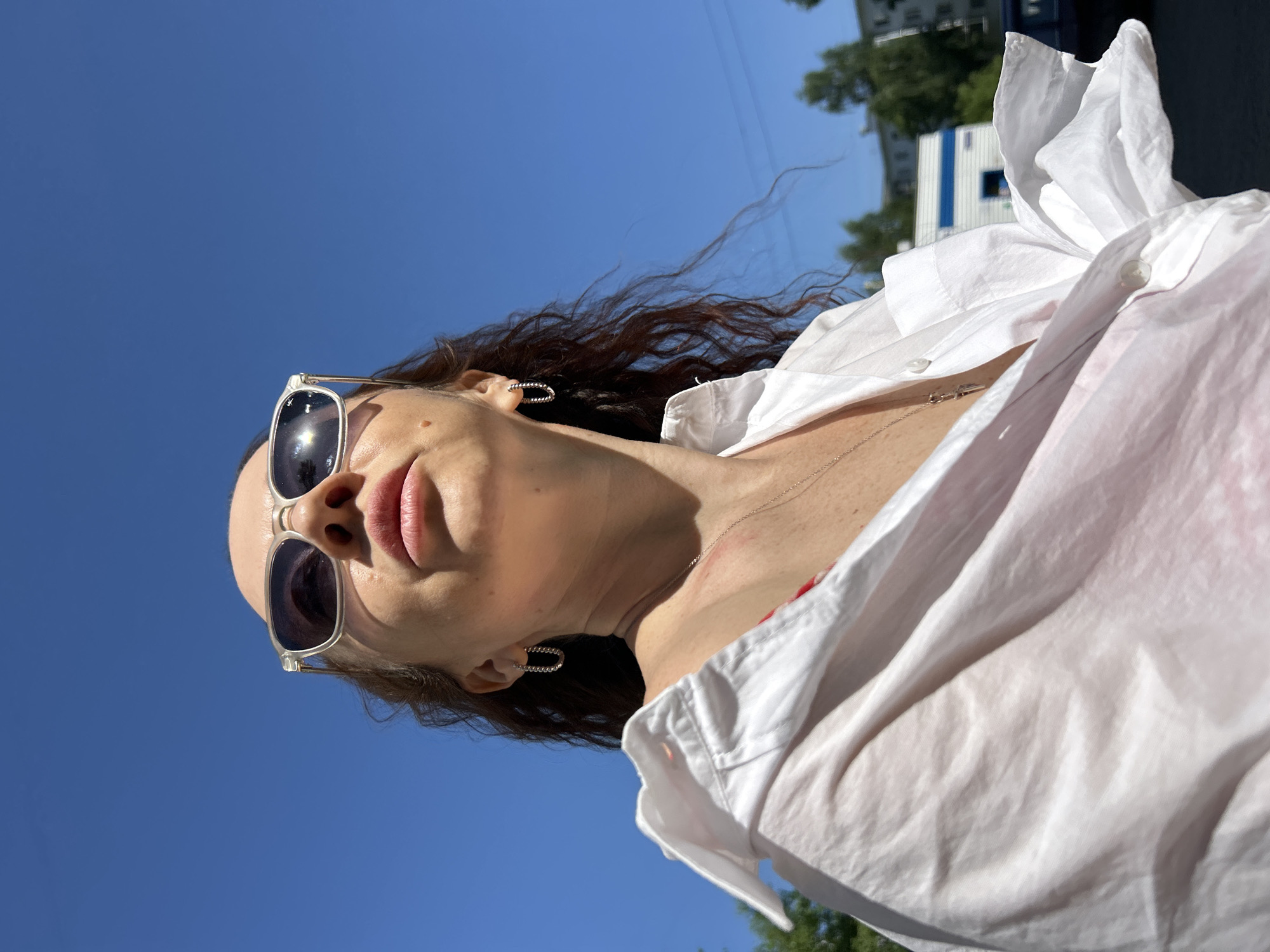 A low-angle outdoor shot shows a woman with long, curly brown hair, wearing sunglasses and a white button-down shirt. She has light skin and is wearing hoop earrings and a delicate necklace. The background is a clear blue sky. To the right of the woman are trees and a white building with blue accents, indicative of an urban setting. The location is Chaikovsky, Russia.