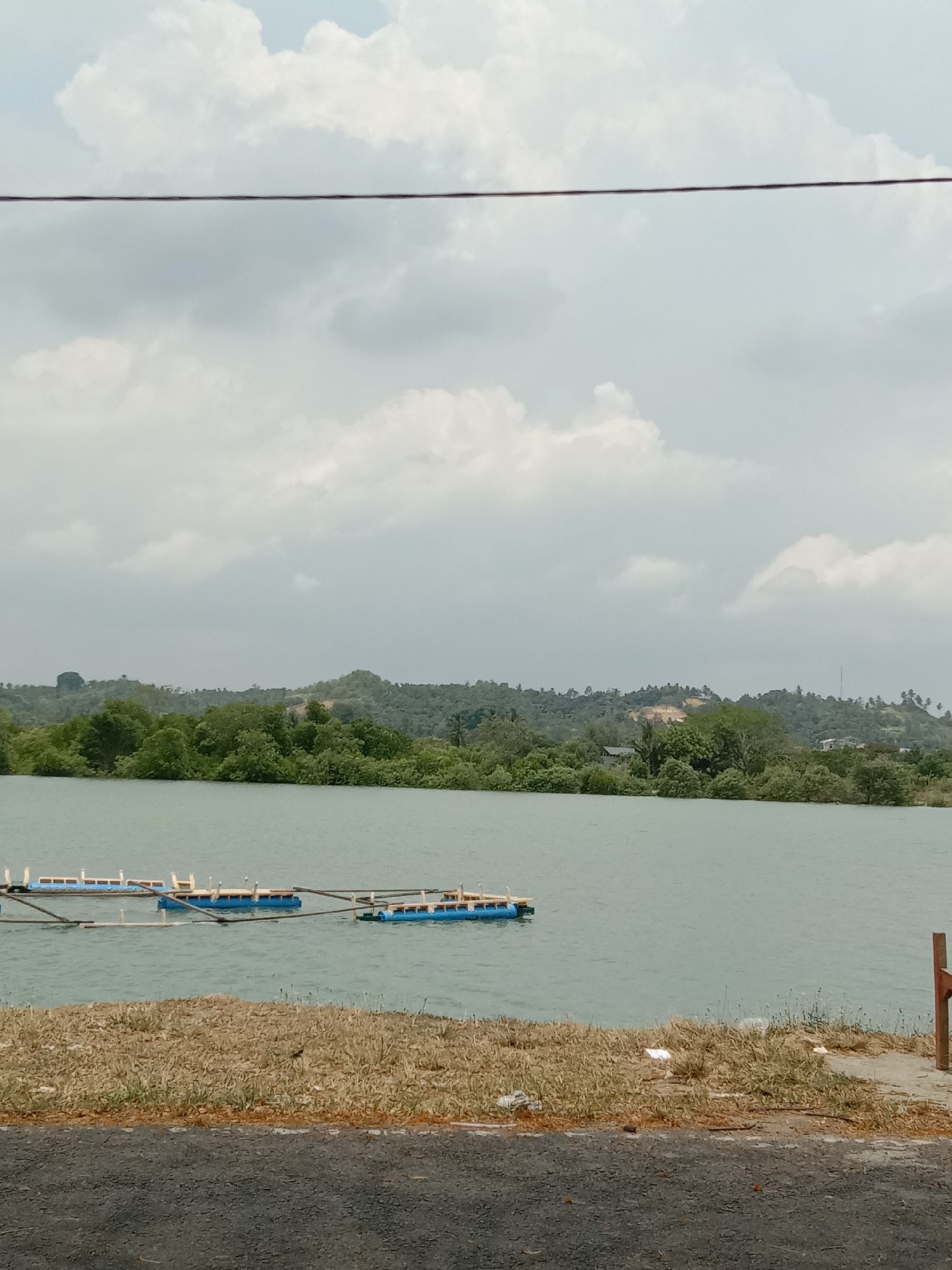 The image depicts a tranquil, overcast day at what appears to be a lake or large pond in Lhokseumawe, Indonesia. In the foreground, there's a stretch of dry, yellow-brown grass bordering a paved road. In the water, there are several blue floats with a supporting structure above them, likely part of a floating platform or aquaculture setup. Across the water, a lush green hillside rises with a variety of trees and some visible structures, indicating a settled area. The sky is dominated by gray and white clouds, suggesting a potentially wet or overcast weather pattern. A single electrical wire runs across the upper part of the frame. The general setting suggests a rural or semi-rural location within the city limits, near a body of water utilized for some sort of commercial or recreational activity. There are pieces of trash, such as white plastic, scattered on the grass at the edge of the water.