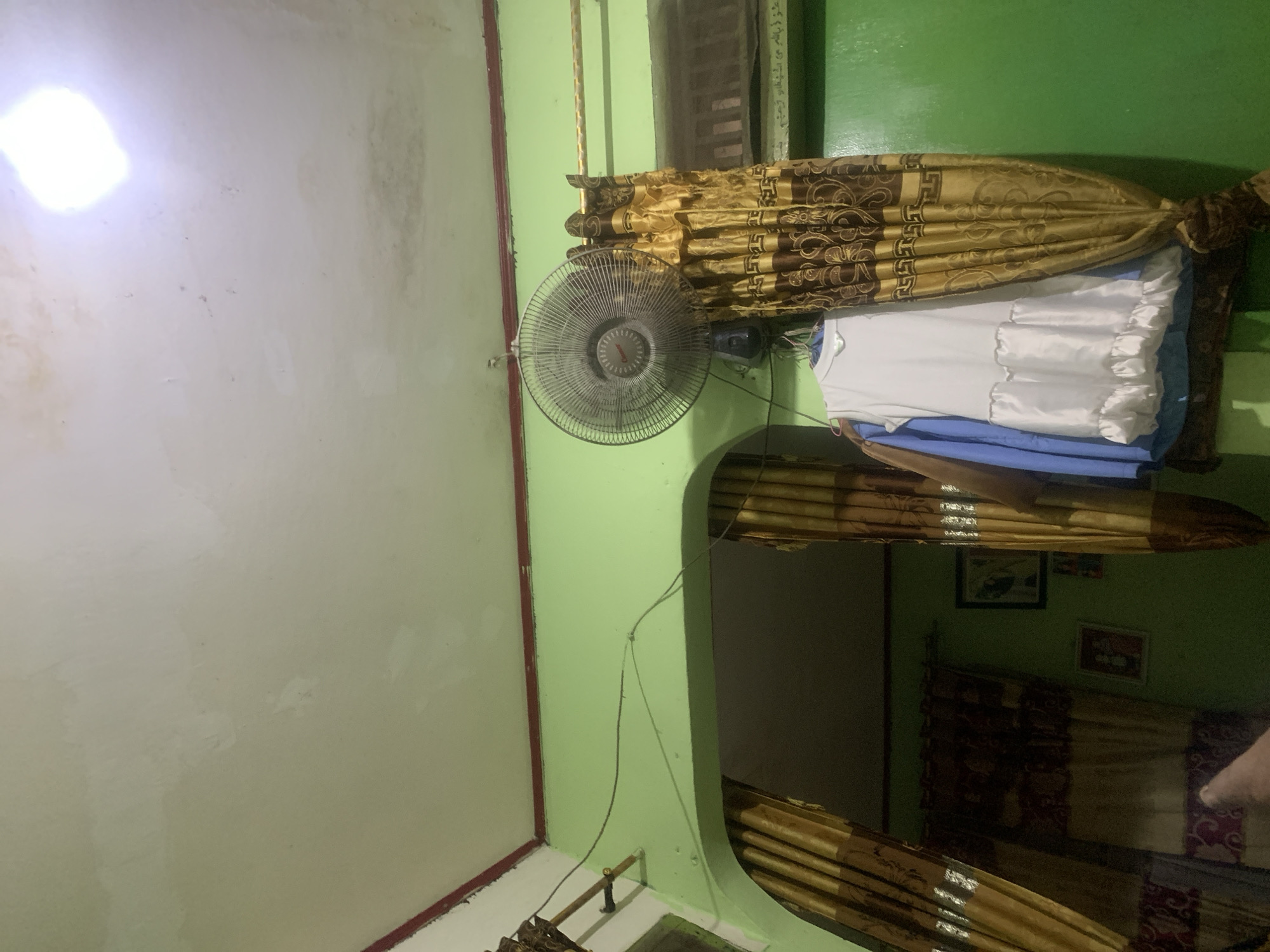 The image depicts an interior scene, likely a residential room, in Palembang, Indonesia. The walls are painted light green, and the ceiling appears to be textured and shows some staining or water damage, particularly near a bright light source at the top right of the frame.
Dominating the upper center of the image is a wall-mounted electric fan, its oscillating head visible. Below the fan, a gold and brown patterned curtain is drawn, partially obscuring the view. To the left of the curtain, hanging on a clothes hanger, is a white dress with ruffled details at the hem. Behind the white dress, a blue garment is visible.
The room features an arched doorway on the left, leading to a darker area where more gold and brown curtains are present. On the green wall to the right, there are at least two framed pictures.
The lighting is dim overall, with a bright light source directly overhead, suggesting it might be nighttime or a room with limited natural light. The overall impression is of a simple, lived-in domestic space.