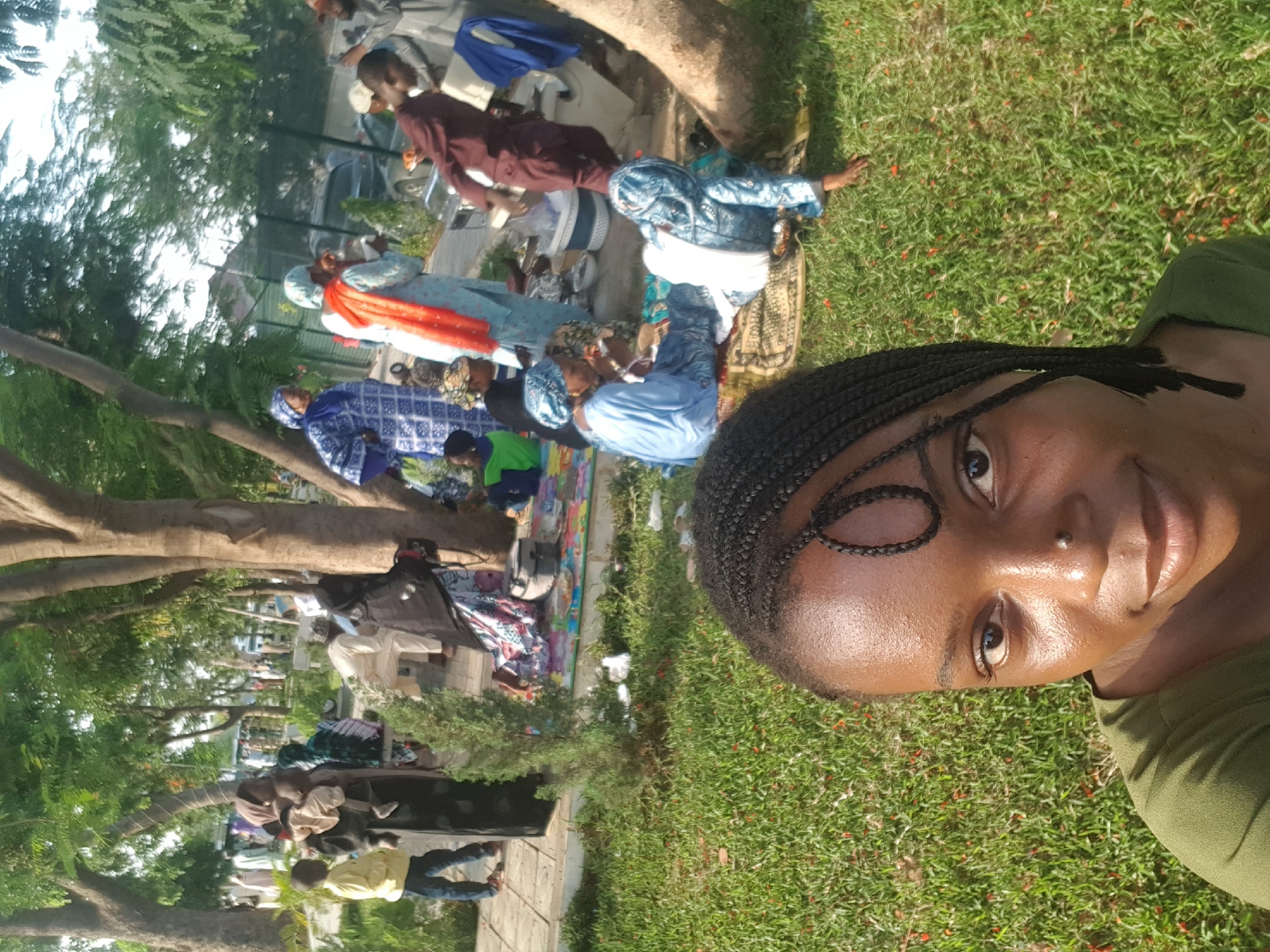 A woman is taking a selfie on green grass. In the background, people are gathered outdoors under trees. The location is identified as Bauchi, Nigeria. People are wearing various clothes and some are sitting on the ground, while others are walking. There are also trees and a partially visible building in the background.