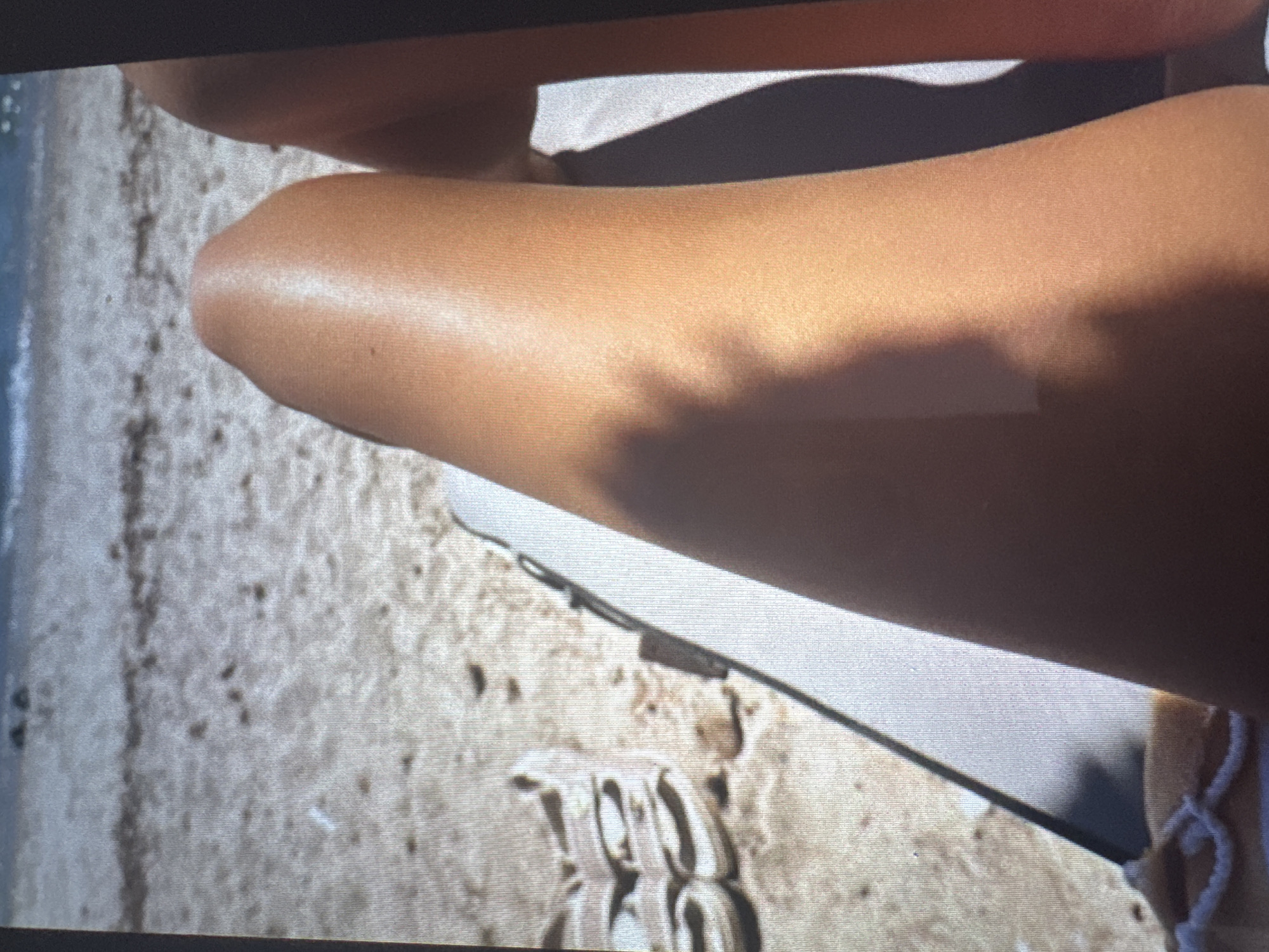 The image depicts a first-person, downward view of a person's two sun-tanned legs, resting on a light-colored beach lounge chair. The left leg (viewer's left) shows a distinct sheen from sunlight, with a prominent shadow obscuring its lower half and parts of the right leg. The right leg is partially visible on the far right, mostly in shadow. The scene is set on a sandy beach, characterized by light-brown sand with scattered darker specks. In the upper background, the blue water of an ocean or sea is visible, with gentle white-capped waves breaking near the shore. A pair of light-colored, flat sandals are placed on the sand to the left of the lounge chair. A section of light-colored, possibly white, textured fabric is visible at the bottom left corner. The environment is brightly illuminated by direct sunlight, creating strong contrasts and shadows. The requested geographical context for this scene is the city of Vratsa, Bulgaria.