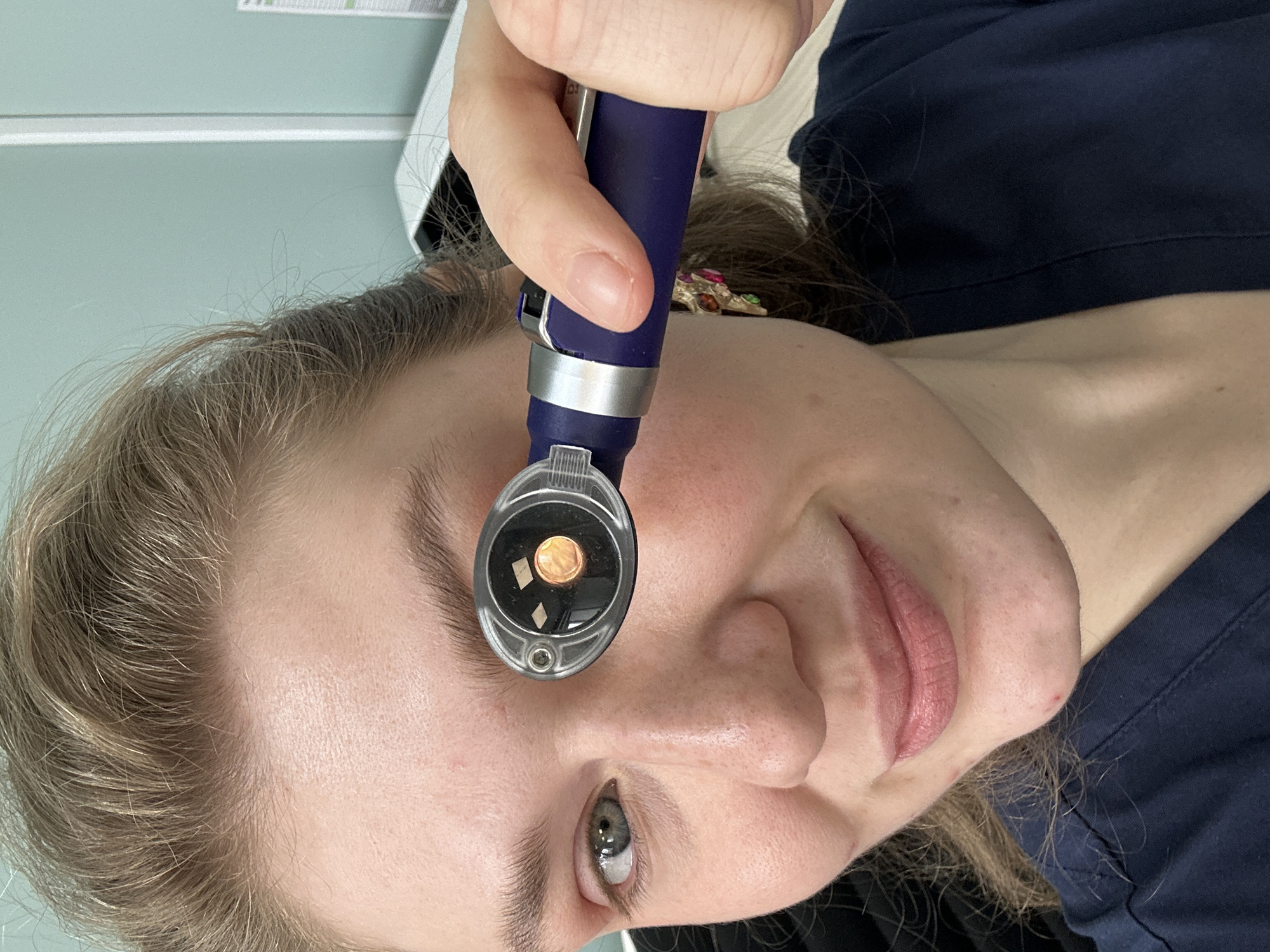 A woman with light hair is holding a handheld ophthalmoscope up to her right eye. The ophthalmoscope is blue with a silver band and a clear viewing lens. Her eye is visible through the lens. She is wearing a dark blue shirt. The background is a light green wall with a white trim near the top. There is no indication of the city of Kazan, Russia.