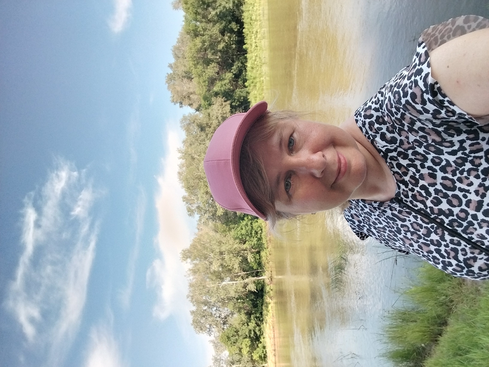 In this selfie, a woman with blonde hair and fair skin, wearing a pink baseball cap and a leopard-print blouse, is positioned in the foreground, looking towards the camera with a slight smile. She is situated beside a calm body of water, likely a river, with lush green trees and vegetation lining the opposite bank. The sky above is a clear blue with scattered wispy white clouds, indicating fair weather. The background features a dense treeline and a gentle ripple on the water's surface, suggesting a natural, serene environment. The location is implied to be the city of Chkalov, Russia, given the context.