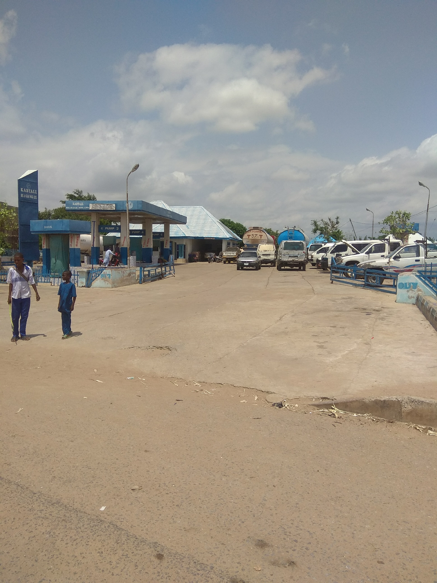This media file depicts an outdoor scene at a gas station. Two young boys, one in a white shirt and blue pants, the other in a blue outfit, are walking in the foreground. The gas station itself features a blue sign with the text "KABTALL OIL & GAS NIG. LTD." A covered structure with pumps is visible, labeled with "DIESEL" and "PUMP 1", "PUMP 2", and "PUMP 6". Several vehicles, including trucks and cars, are parked at the station. One truck has the text "T 57 T" on its side. The scene is set on a paved area under a bright, partly cloudy sky, suggesting a daytime setting. The environment appears to be a semi-arid or dusty region, given the ground cover and dry vegetation scattered around. The overall atmosphere is one of everyday activity at a commercial establishment.