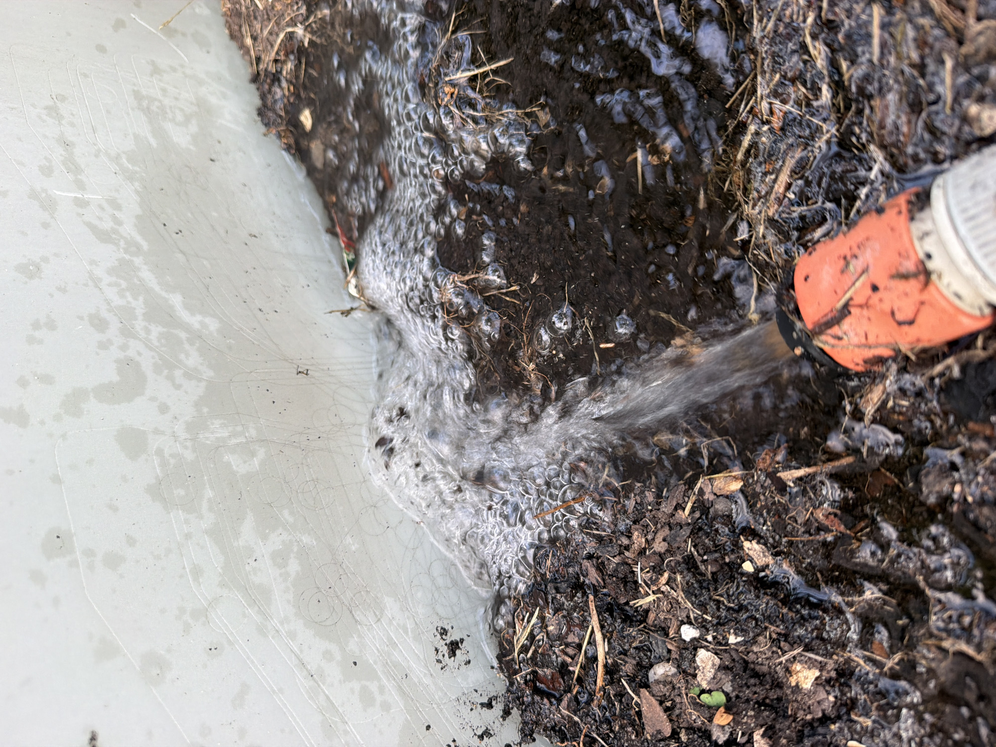 A garden hose with an orange nozzle is spraying water into a pile of compost. The compost is a mixture of dark soil, wood chips, and dried plant matter. The water is creating bubbles and ripples as it hits the compost. The scene appears to be outdoors, likely in a garden or yard, given the presence of compost and a garden hose. There is no indication of the specific location within Bayreuth, Germany, as the image provides no landmarks or identifying features of that city.