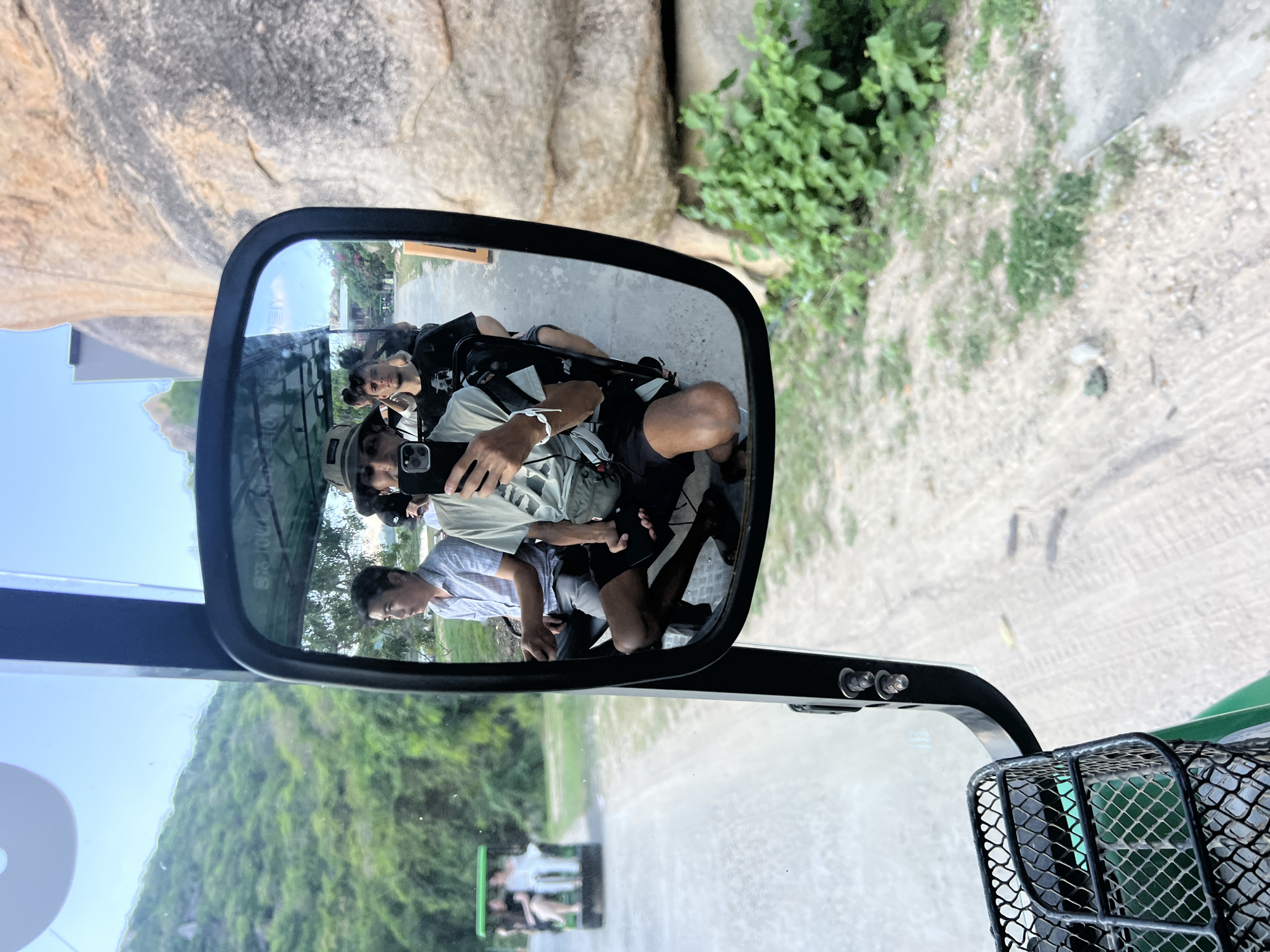 A mirror-image selfie is taken from the perspective of a passenger in a golf cart, capturing the scene ahead and to the side. The passenger, wearing a hat and a light-colored t-shirt, holds a phone to take the photo. Beside them, another passenger in a black t-shirt is visible. In the background, reflected in the mirror, a man in a light blue patterned shirt is seated, and other passengers are seen in the cart. Further ahead, another golf cart with passengers is driving on a dirt road. The environment features lush green hills on the left and a large rock formation on the right, suggesting a natural or park-like setting. The scene is likely set in Vĩnh Hải, Vietnam, given the context of the location.