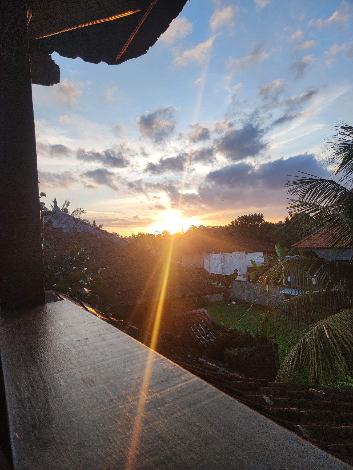 The image depicts a sunset over a residential area in Ubud District, Indonesia. The sun is positioned low on the horizon, with rays of light shining through clouds and across the landscape. Several buildings with tiled roofs are visible amidst trees and vegetation. The foreground includes a wooden ledge or railing, providing a perspective from an elevated location.