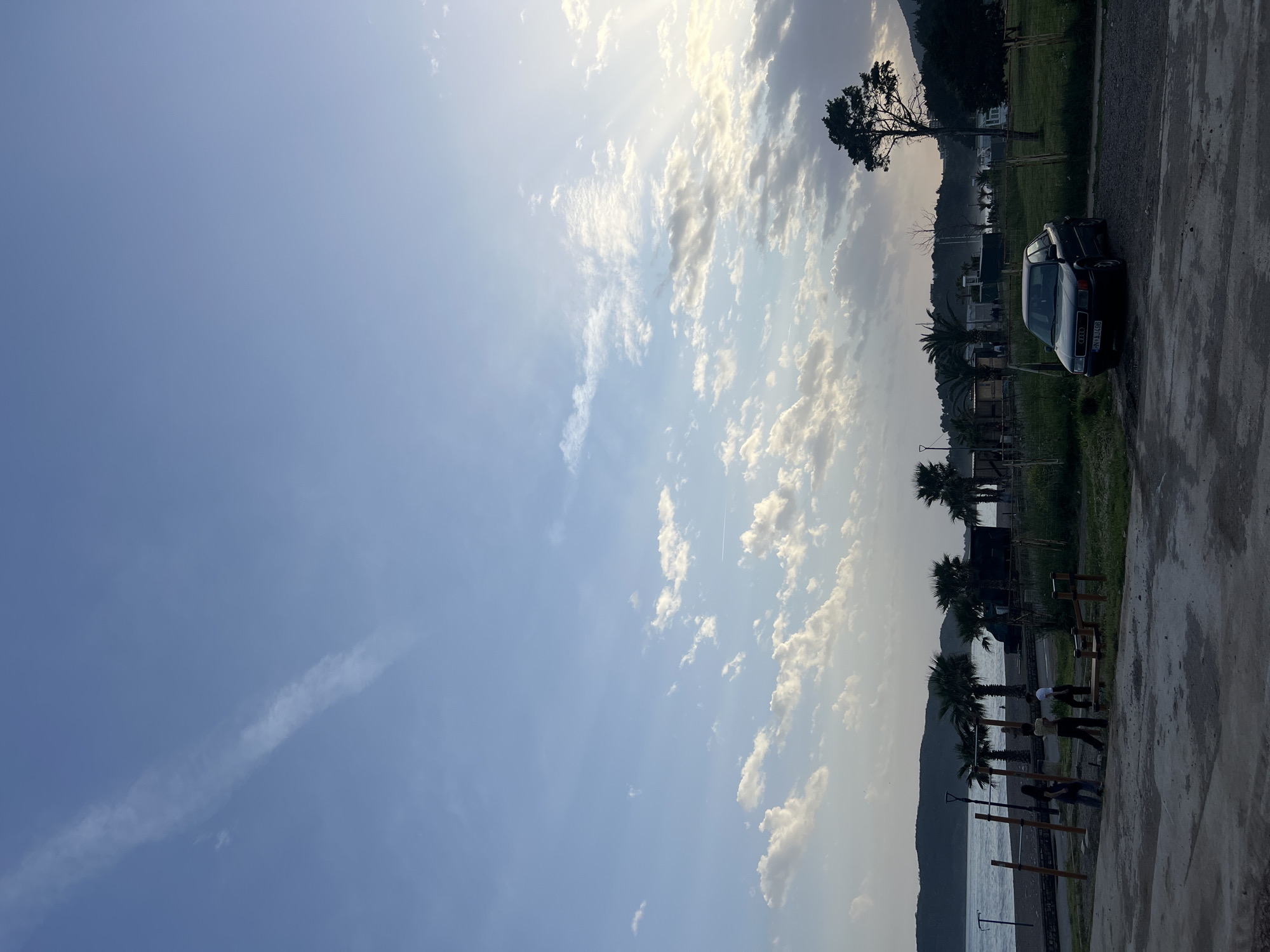 The image depicts a coastal scene in Boreti, Montenegro, under a partly cloudy sky. The leftmost half of the image is dominated by the blue sky interspersed with wispy and puffy clouds. On the right side, the terrain transitions from a concrete path in the foreground to a grassy area with palm trees, exercise equipment, and a parked black car. People are visible using the exercise equipment near the shoreline. The background includes a coastline with mountains in the distance.