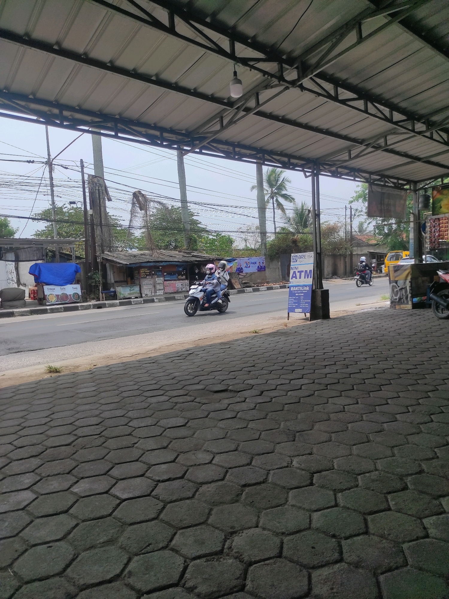 The image captures a street scene in Cipatat, Indonesia, viewed from under a covered walkway. In the foreground, a textured gray hexagonal paver surface leads towards a paved road. The road has a distinct curb separating it from the sidewalk.
On the road, two people on a white scooter are moving from left to right. The scooter rider is wearing a blue jacket and a white helmet, while the passenger wears a pink helmet and a light-colored top.
Across the street, a row of modest buildings and stalls line the roadside. One stall on the left has a blue tarp covering goods and a sign with images of food items like "Tempe", "Sate", and "Nasi". Further down, a small building with a corrugated metal roof has various posters and signs, including one for "BRI Link Mini ATM". Another yellow vehicle is visible in the background, and a portion of another motorcycle is parked on the right side of the image.
The environment is characterized by overhead wires, utility poles, and trees, including palm trees, suggesting a tropical or subtropical climate. The sky is overcast, and the overall lighting suggests it is daytime. The structure providing the covered viewpoint has a metal roof supported by a metal frame.