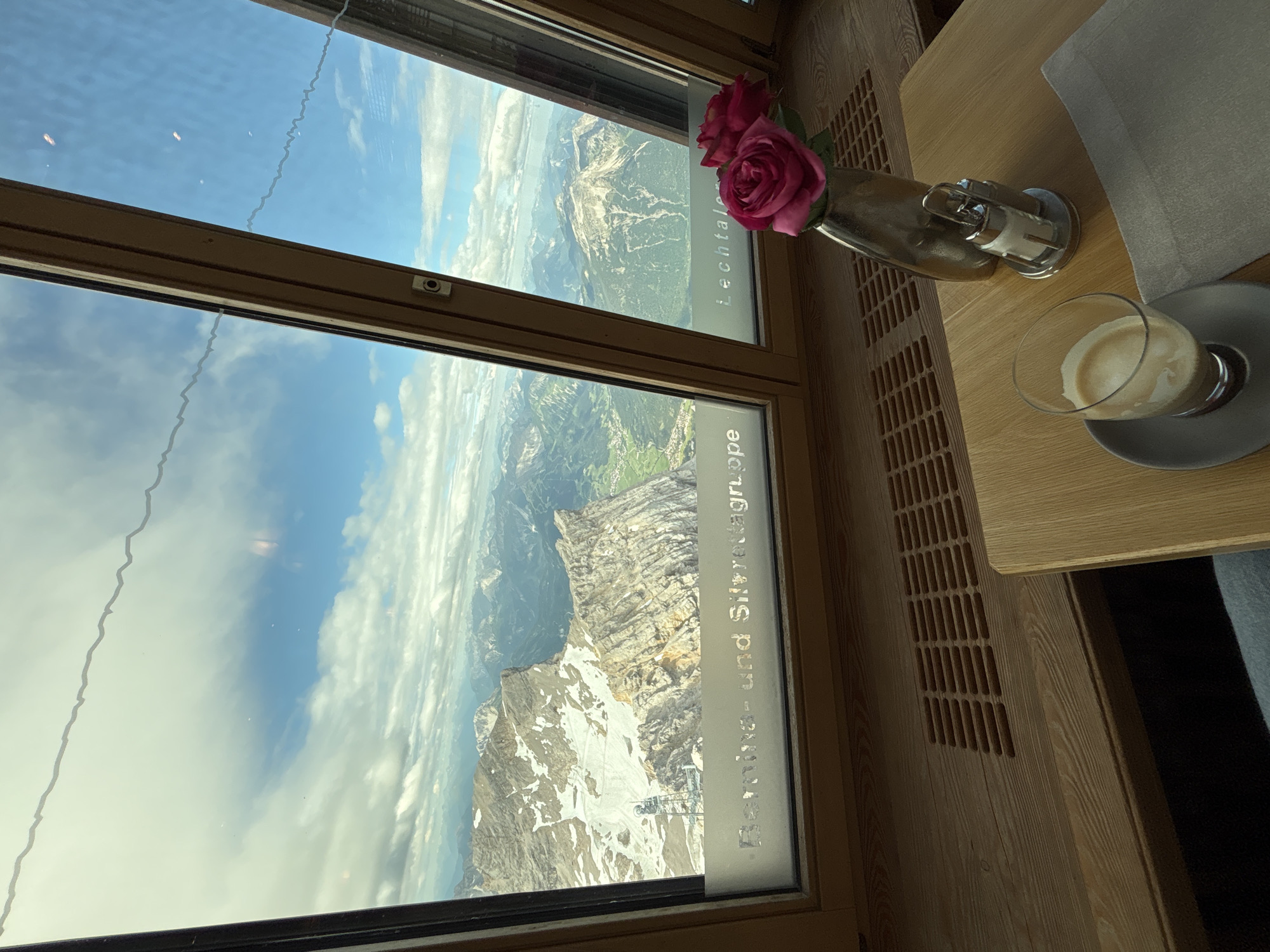 The photograph is taken from inside a building with a large window offering a panoramic view of a mountainous landscape. The window is framed by wooden beams. Through the window, a rugged mountain range with snow-capped peaks and green valleys is visible under a cloudy sky. In the foreground, on a wooden table, is a glass of coffee with crema, a salt and pepper shaker, and a vase with two pink roses. A grey napkin is placed next to the coffee. The text "Bernina- und Silvrettagruppe" and "Lechtaler" is visible etched on the lower part of the window, indicating a location in the Alps, likely near Ehrwald, Austria, given the reference to the Lechtal valley. The scene appears to be a serene moment in a cafe or restaurant with a stunning natural vista.