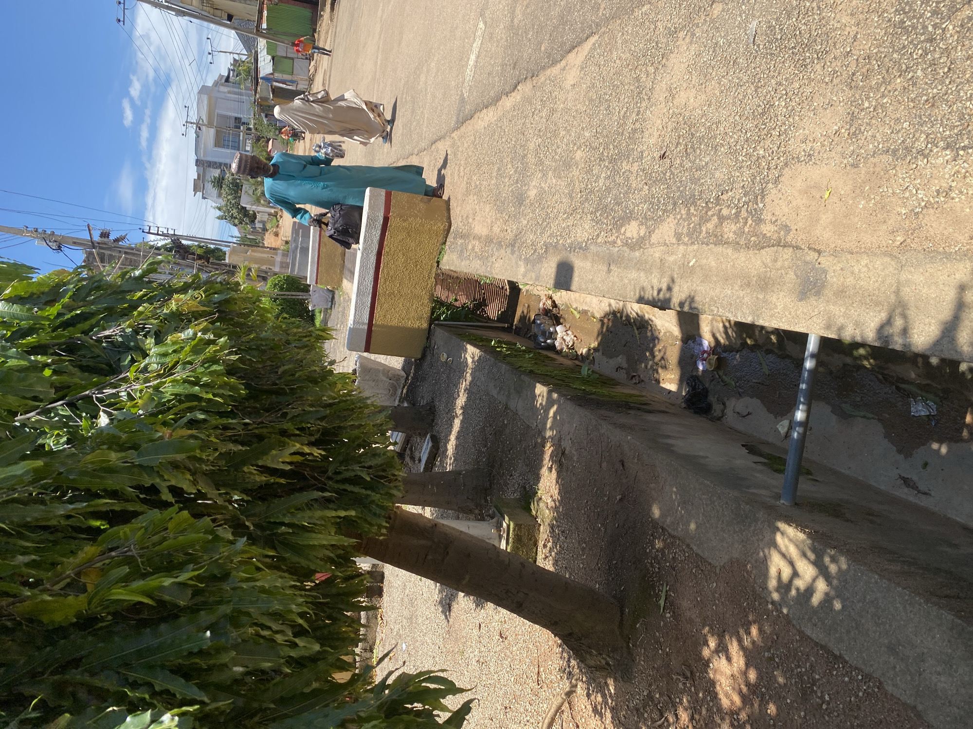 The image captures a sunny day in Jos, Nigeria, depicting a street scene with a focus on a drainage ditch and passersby. In the foreground, a deep concrete drainage ditch runs along the side of a paved road, filled with murky water and scattered debris. A metal pipe extends from the side of the ditch into the road. Adjacent to the ditch, a rough, gravelly area is partially shaded by the dense foliage of a large tree with long, drooping leaves.
On the road, a man wearing a blue robe and a patterned cap walks towards the viewer, carrying a black bag and some items in his other hand. Behind him, a woman in a beige hijab and robe also walks away from the viewer. Further down the road, two other figures are visible: one person in a red shirt and another on a motorcycle. In the background, modern buildings and power lines are interspersed with smaller structures, indicating a developed urban environment. The sky is bright blue with scattered white clouds.