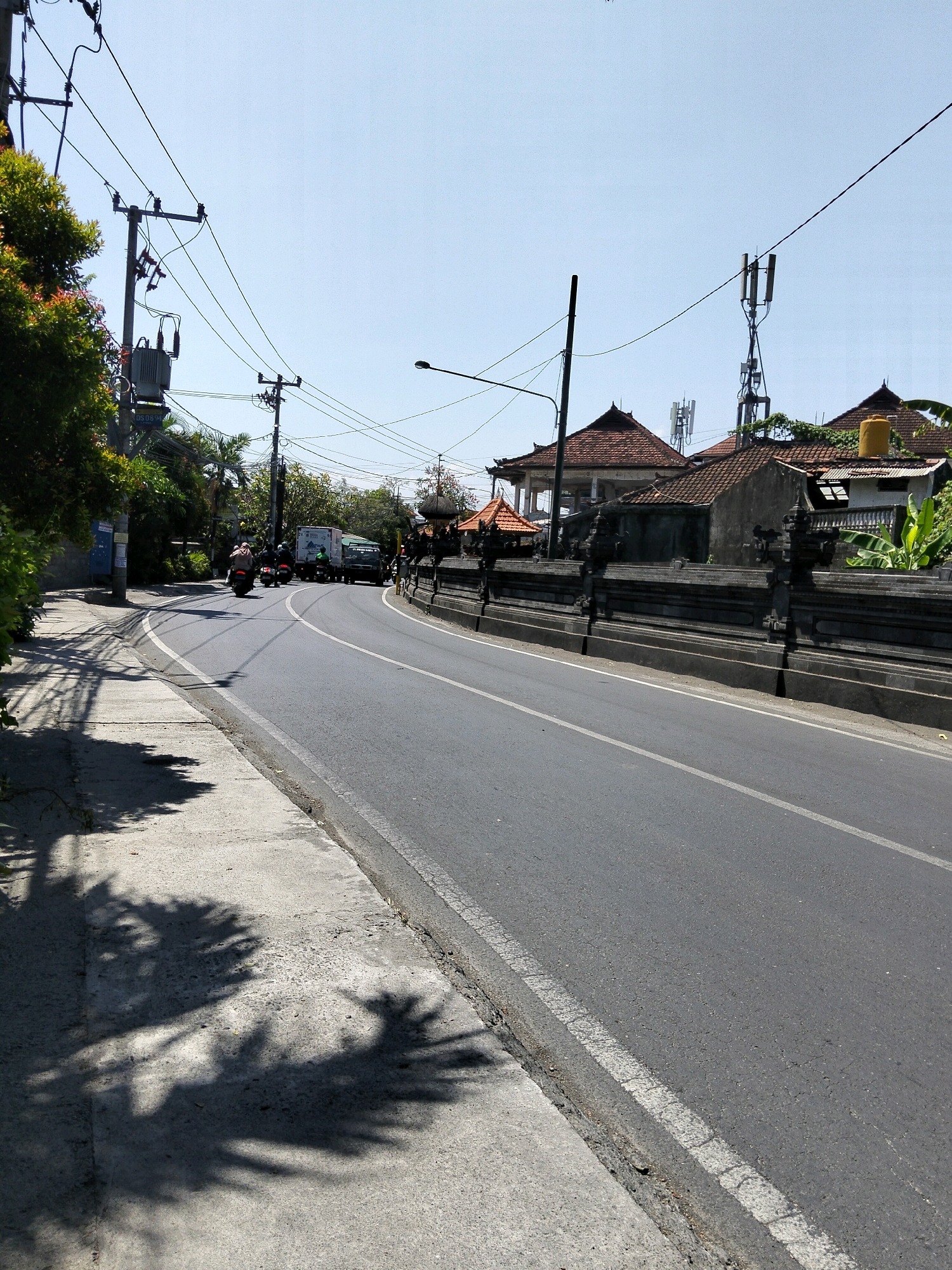 A paved road with a white dashed line runs through a tropical environment in Denpasar, Indonesia. The road is bordered by a sidewalk with palm tree shadows and a stone wall with ornate pillars. Along the road, there are several motorbikes and a truck. Buildings with tiled roofs and cell towers are visible in the background. The sky is clear and blue.