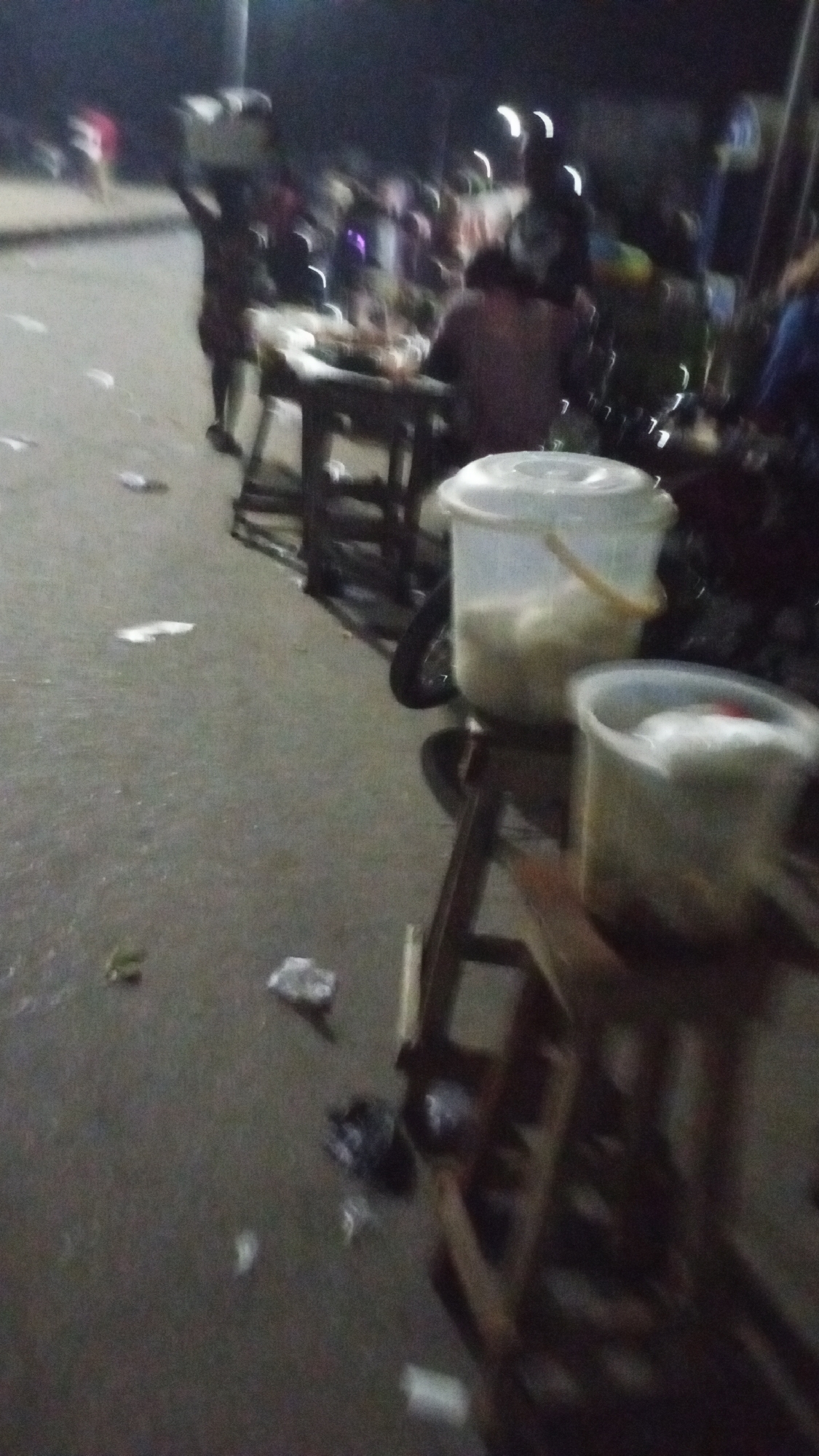 The image shows a street scene in Lokoja, Nigeria, at night. The lighting is dim and possibly from overhead street lamps. There is a gathering of people, some seated at tables. One individual is carrying a load on their head. Two plastic buckets are situated on wooden stools in the foreground. Trash litters the street. There is a motorcycle parked near the tables.
