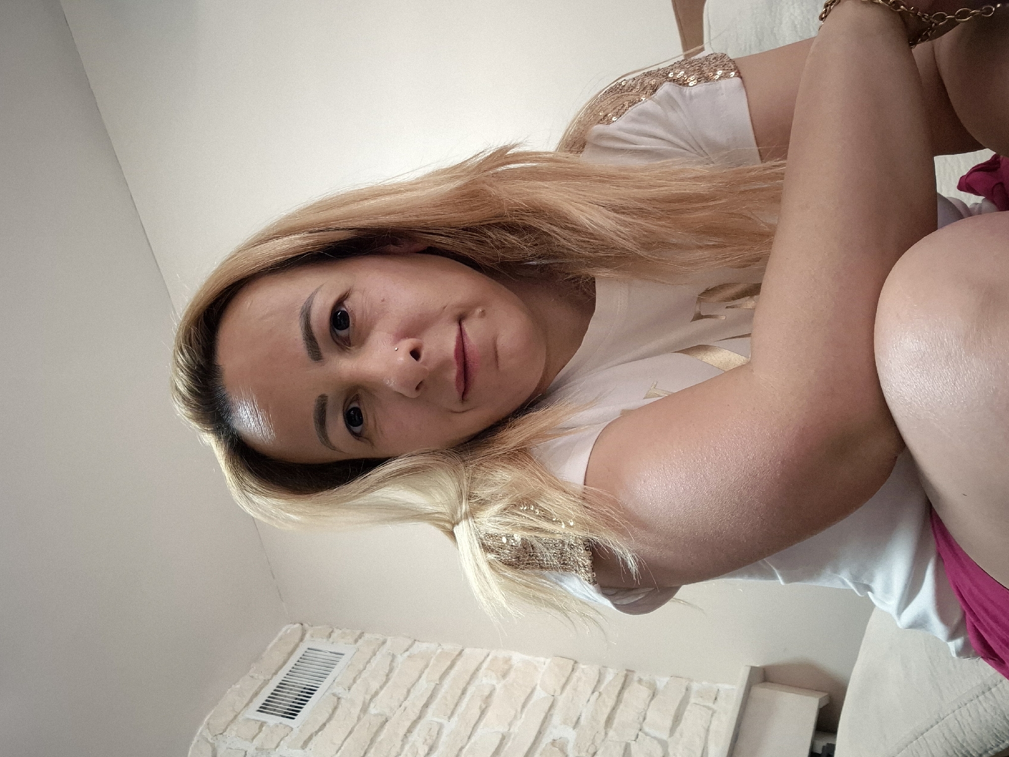 A medium shot captures a woman with blonde hair and fair skin, sitting indoors and looking towards the camera. She has a nose piercing and is wearing a white t-shirt with gold embellishments on the shoulders. Her arms are crossed in front of her chest. The background includes a wall with a decorative stone finish and a ventilation grate on the left side, with a white wall and a bed on the right. The lighting is soft and diffused, indicating an indoor setting.
