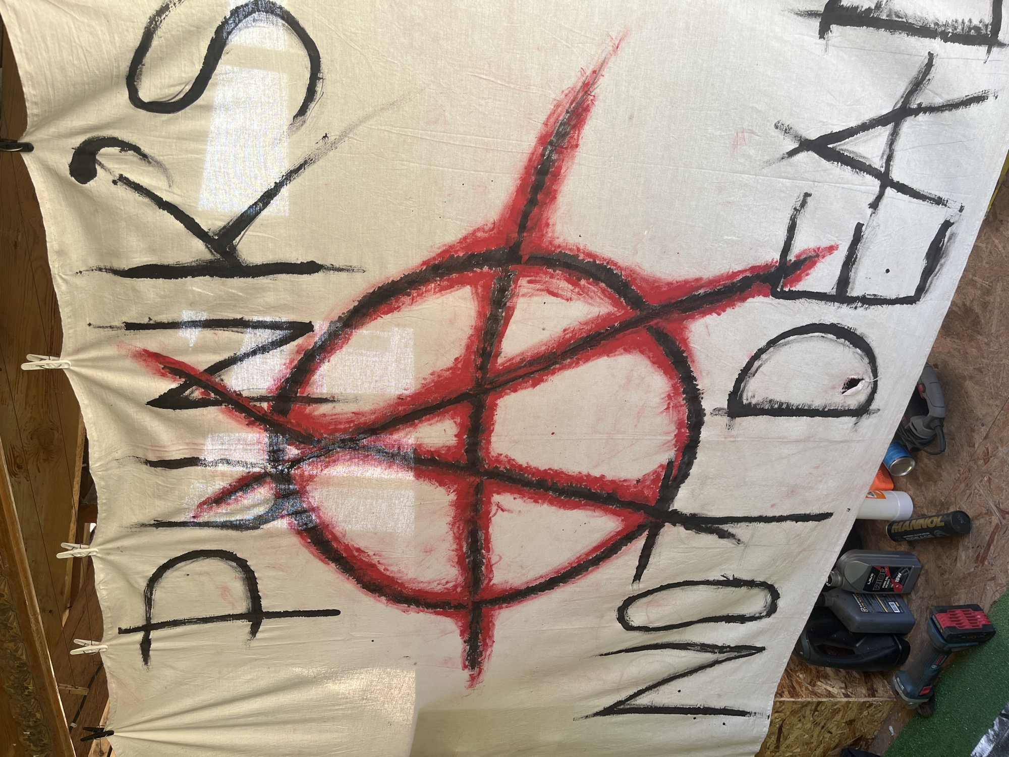 A white cloth banner with hand-painted black and red anarchist symbols and text hangs indoors. The text reads "PUNKS NOT DEAD". The anarchist symbol consists of a red circle with a black outline encompassing a red "A" that extends beyond the circle's perimeter. The cloth is hung using clips, and the background shows a wooden structure, potentially a workshop or shed. Tools and containers, including power tools and oil bottles, are visible on the right side of the image.