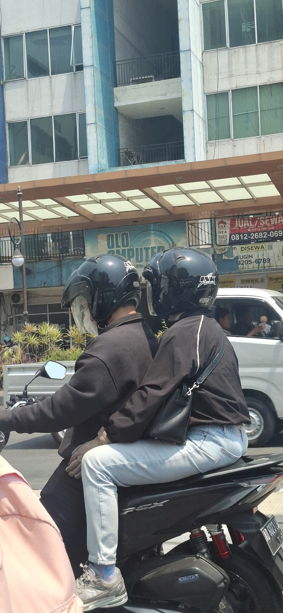 A medium shot captures two individuals wearing black helmets and dark tops, riding a black scooter on a sunny day in Surakarta, Indonesia. The rider in front has a hand resting on the pillion rider's lap, and the pillion rider's arms are around the rider. Both are wearing light-colored jeans. The pillion rider has a black crossbody bag. In the background, a white car is visible, and beyond that, a multi-story building with light-colored facades and blue accents, featuring rows of windows, stands out. A banner with text, including "OLO COMPUTER," and other advertisements is displayed across the front of a commercial building. The scooter's license plate is partially visible. The overall setting appears to be a street in a commercial area of the city.