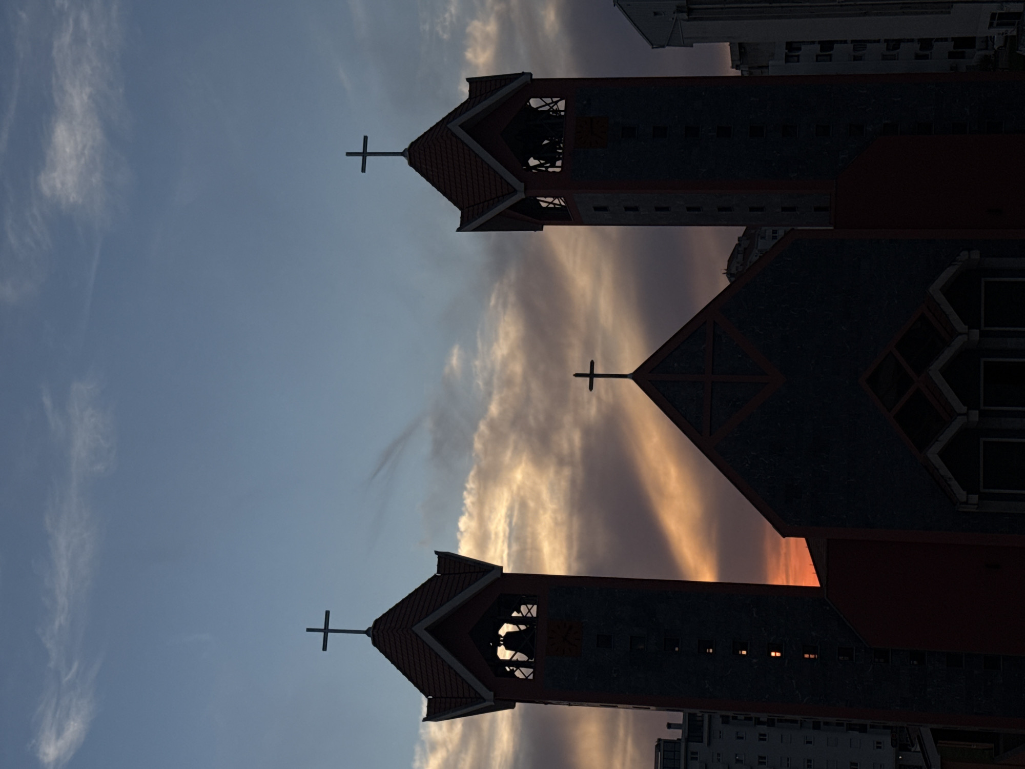 The image depicts a church or cathedral in Bar, Montenegro, viewed from a low angle. The camera is oriented upward, capturing the silhouette of the building against the sky. The sky transitions from blue to orange/yellow, potentially indicating sunrise or sunset. The image includes two towers, each topped with a cross. The church structure appears dark, allowing details like the rooftop, window openings, and decorative features to stand out against the bright sky. There are tall buildings adjacent to the church.
