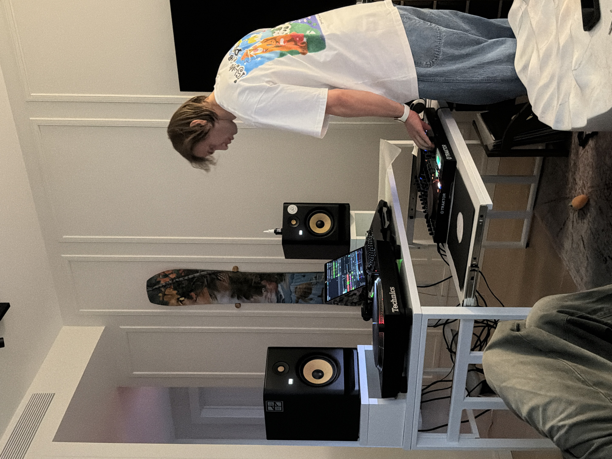 A young man is intensely focused on a DJ setup within what appears to be a home studio in Moscow, Russia. He has light brown hair and is wearing a white t-shirt with a colorful graphic print featuring cartoon characters, paired with light blue jeans. He leans over a white, minimalist table, operating a black DJ mixer marked with "TRAKTOR" logos. A white wristwatch is visible on his left wrist as he adjusts the controls.
The DJ station includes two black studio monitor speakers with distinctive yellow cones, bearing "KRK" and "NI" (Native Instruments) branding. They are positioned on the white table, flanking the central equipment. To the left, a black turntable or media player is visible, clearly labeled "Technics," with a tablet or laptop displaying audio waveforms and controls beside it. Cables are visible underneath and behind the setup, indicating active use.
In the background, leaning vertically against a light-colored wall with decorative molding, is a snowboard featuring a detailed camouflage pattern with animal faces, possibly wolves or foxes. On the floor to the right of the table, a small, orange, oval-shaped object is noticeable. The lighting is bright and even, typical of an indoor, well-lit environment. The scene depicts a dedicated space for music creation or performance.