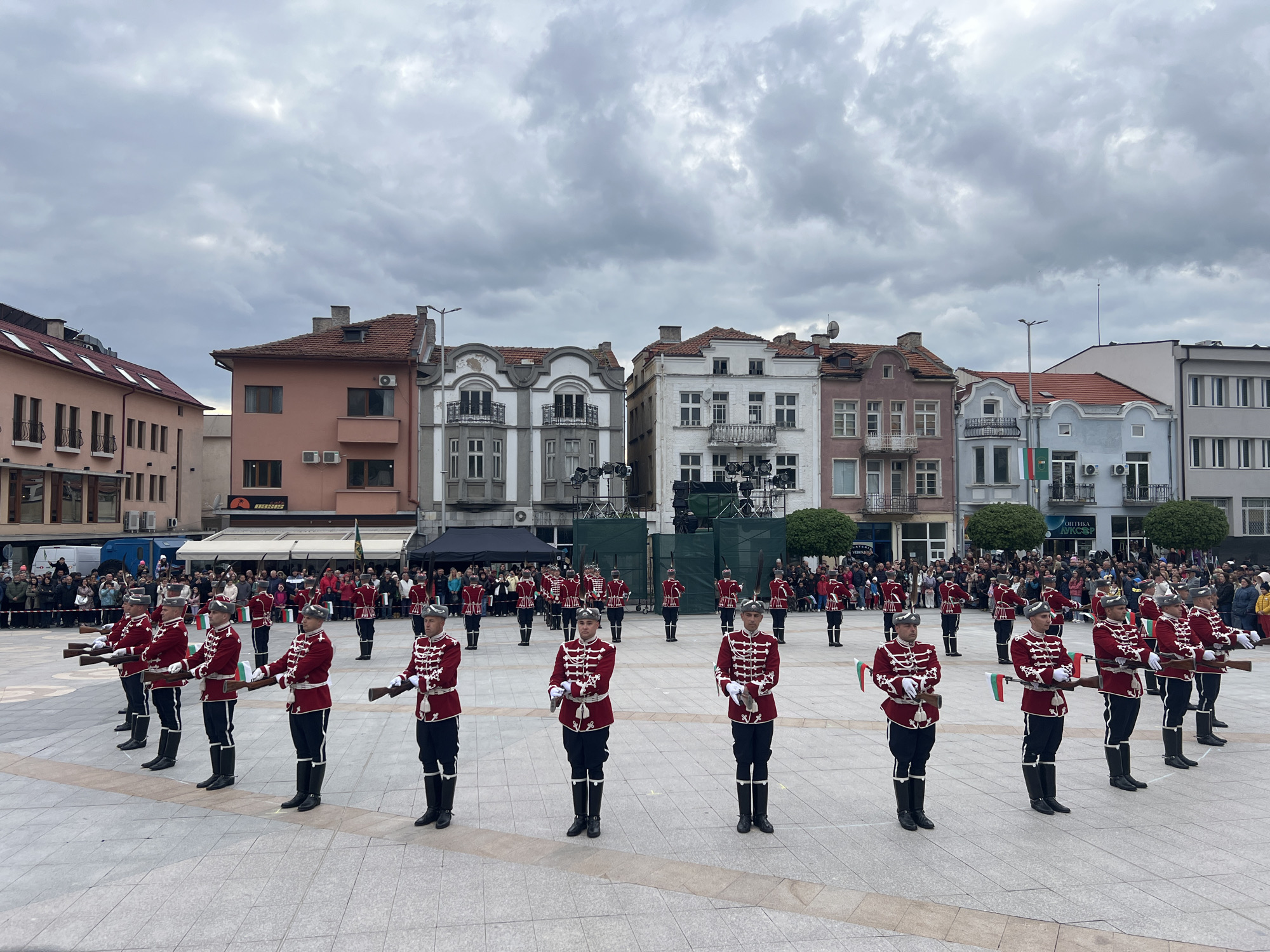 This media file captures a formal ceremonial event held in a town square in Panagyurishte, Bulgaria. The central focus is a large group of uniformed guards, likely a military or honor guard unit, engaged in a drill or parade.
The guards are distinctive in their uniforms, which feature dark red jackets adorned with intricate white and gold braiding, dark trousers, and tall black boots. They wear unique caps and most carry rifles, many of which are decorated with small Bulgarian flags. They are arranged in two curved lines, facing inwards towards a central aisle on the paved square, performing synchronized movements.
Behind the guards, a substantial crowd of civilians stands observing the event from behind barriers, suggesting a public celebration or commemoration. In the background, the square is framed by a collection of charming, multi-story buildings displaying traditional European architectural styles. These structures feature varied facades in colors such as pink, white, grey, and light blue, often including balconies and red-tiled roofs. In the mid-ground, a temporary stage or setup with lighting equipment is visible.
The event occurs during the daytime under a heavily overcast and cloudy sky, which provides soft, diffuse lighting. Visible text on commercial signs includes "OASIS" and "ОПТИКА АКСАР" (Optics Aksar) in Cyrillic script. The Bulgarian flags and the Cyrillic text confirm the specified location. The scene conveys a sense of formal public gathering and national pride.