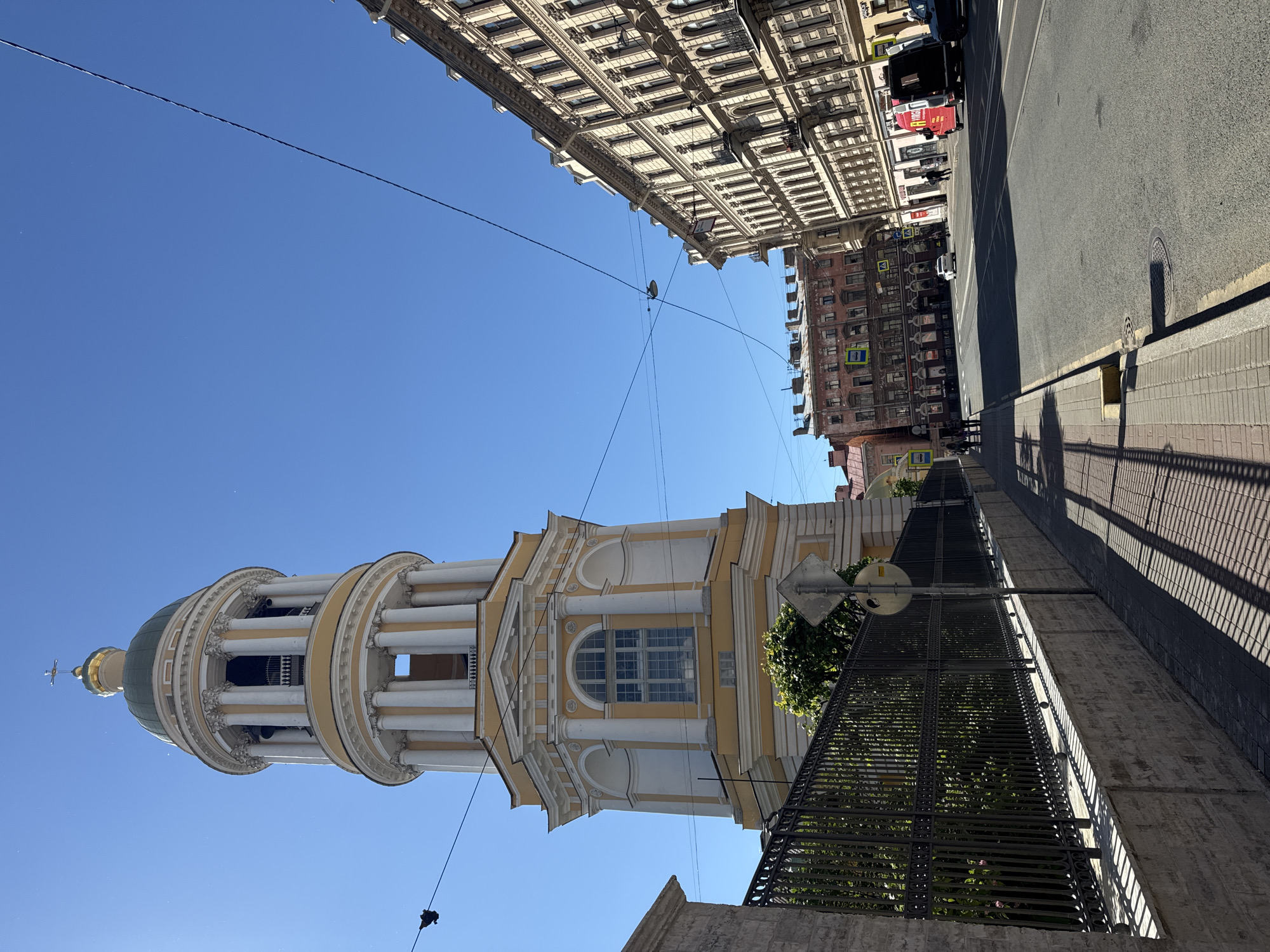 This is a street-level, eye-level, medium-shot, outdoor, daytime photograph taken on a clear day in Saint Petersburg, Russia. The image captures a view looking down a street with several buildings and a tall church bell tower. The bell tower, which is yellow and white with a green dome and a cross on top, dominates the left side of the frame. It has multiple tiers with columns and arched openings. A black metal fence with a stone base runs along the sidewalk in front of the tower. To the right of the bell tower, there are several multi-story buildings lining the street, typical of Saint Petersburg's architecture, with light-colored facades and numerous windows. The street itself is paved and has lane markings. There are a few cars and a red van visible on the street. A few pedestrians are walking on the sidewalk. Overhead, there are power lines crisscrossing the sky. The overall scene depicts a typical urban environment in Saint Petersburg on a sunny day.
