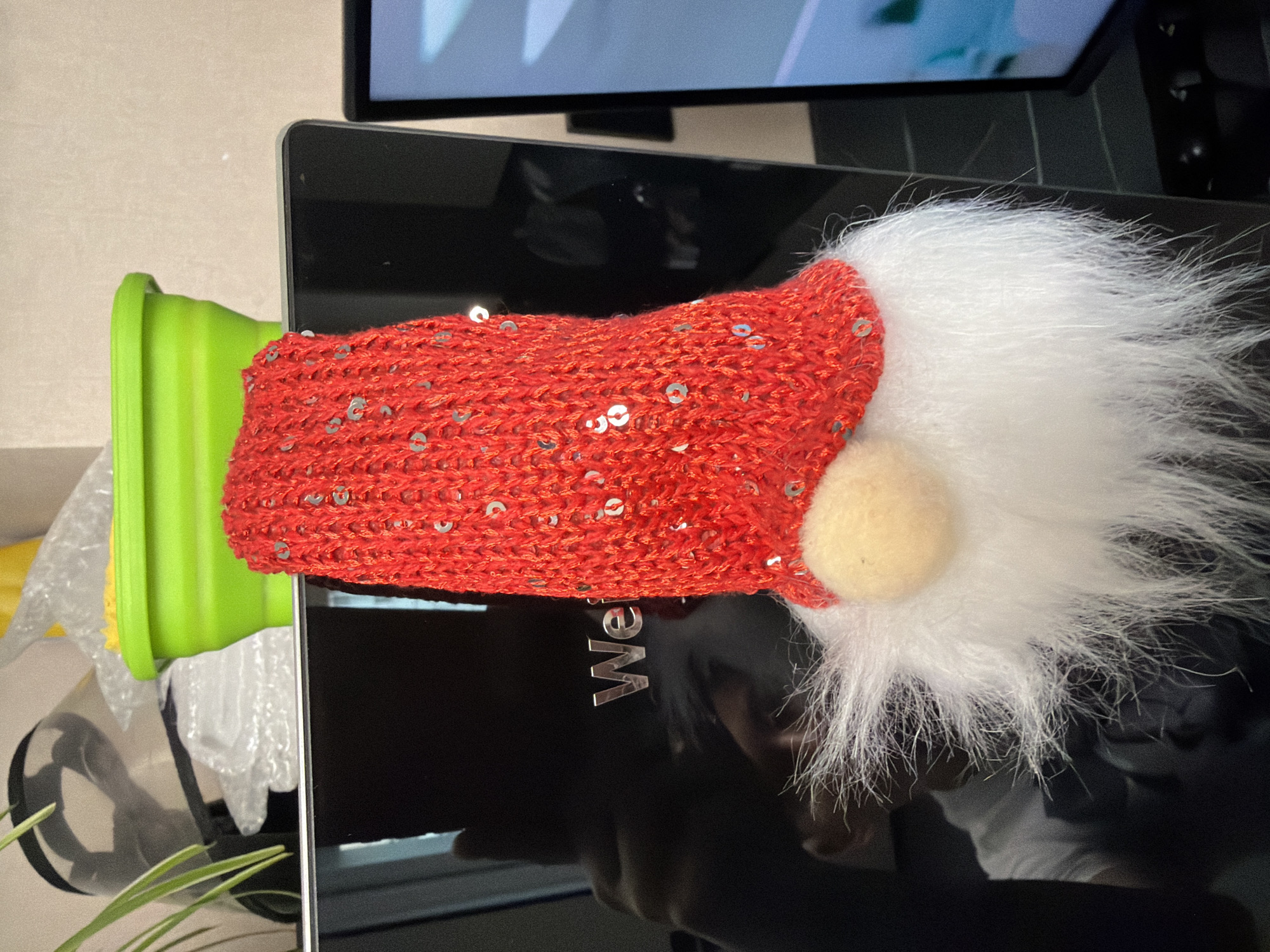 A festive gnome ornament, adorned with a red sequined knit hat and a white fluffy beard, is affixed to a dark, reflective surface, possibly a computer monitor. The gnome's nose is a small, tan pom-pom. In the background, a bright green collapsible bowl is visible, along with other objects that suggest an indoor setting. The dark surface reflects a person wearing a white t-shirt. The image quality suggests it was taken indoors with artificial lighting. The specific location within Moscow, Russia cannot be determined from the provided image.