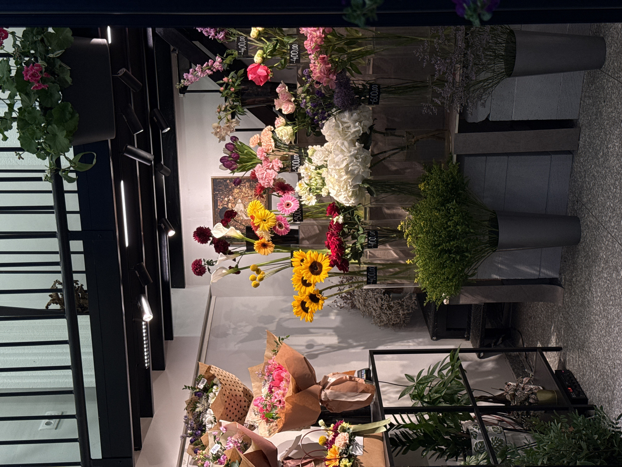 The image depicts an indoor flower shop, likely in Belgrade, Serbia. Various cut flowers are displayed in vases and wrapped bouquets. Flowers include roses, sunflowers, daisies, and other unidentified varieties. Prices are indicated on some stems. The arrangements are displayed along the wall and on shelves. The setting is a modern, well-lit retail space.