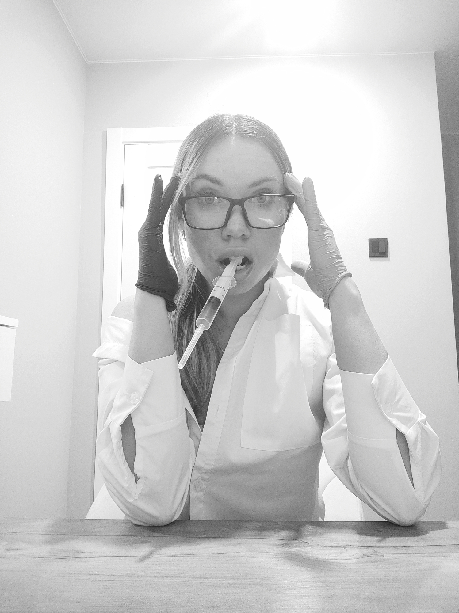 A black and white, well-lit portrait features an adult woman seated at a light-colored wooden table, looking directly at the camera. She wears eyeglasses and a white collared shirt or lab coat, with sleeves rolled up or cuffed. Her left hand is covered by a black surgical-style glove, and her right hand by a lighter-colored (possibly white or beige) glove. Both gloved hands are raised, with fingers splayed near her temples, suggesting a pose of intensity or a dramatic gesture.
A large syringe, appearing to contain a reddish-brown liquid, is held in her mouth, positioned as if she is drinking from it or biting it. Text is visible on the barrel of the syringe, but it is too blurry to be deciphered.
The background consists of plain, light-colored walls. A white door frame is visible to the left, and a dark light switch or electrical outlet is mounted on the wall to the right. The room's minimalist decor offers few specific cues beyond suggesting an indoor environment like a clinic, laboratory, or a home with simple furnishings. The strong, direct lighting creates high contrast throughout the image. The woman's expression is serious and direct, aligning with the dramatic staging of the scene. The image's location context is Votkinsk, Russia.