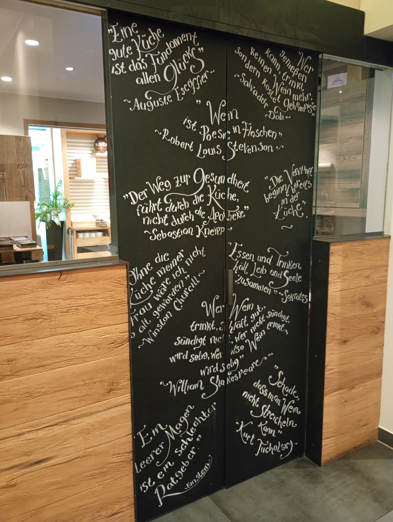 The image shows a black sliding door with white lettering, likely in a restaurant or wine shop. The text consists of quotes about food, wine, and happiness. The door is located in the city of Bad Sachsa, Germany, as indicated by the presence of a sign for "Hotel Stadt Bad Sachsa" in the background.
On the left side of the door, the following quotes are visible:
- "Eine gute Küche ist das Fundament allen Glückes ~ Auguste Escoffier" (A good kitchen is the foundation of all happiness ~ Auguste Escoffier)
- "Wein ist Poesie in Flaschen ~ Robert Louis Stevenson" (Wine is poetry in bottles ~ Robert Louis Stevenson)
- "Der Weg zur Gesundheit führt durch die Küche, nicht durch die Apotheke ~ Sebastian Kneipp" (The path to health leads through the kitchen, not through the pharmacy ~ Sebastian Kneipp)
- "Ohne die Küche meiner Frau wäre ich nie so alt geworden ~ Winston Churchill" (Without my wife's kitchen, I would never have gotten so old ~ Winston Churchill)
- "Ein leerer Magen ist ein schlechter Ratgeber" (An empty stomach is a bad advisor)
On the right side of the door, the following quotes are visible:
- "Wer Wein kann, trinkt keinen mehr, sondern kostet Geheimnisse ~ Salvador Dalí" (He who can, drinks no more wine, but tastes secrets ~ Salvador Dalí)
- "Die Vernunft beginnt bereits in der Küche" (Reason already begins in the kitchen)
- "Essen und Trinken hält Leib und Seele zusammen ~ Sokrates" (Food and drink hold body and soul together ~ Socrates)
- "Wer Wein trinkt, schläft nicht. Wer nicht sündigt, wer nicht trinkt, wird selig, wer also Wein trinkt, wird selig ~ William Shakespeare" (He who drinks wine, does not sleep. He who does not sin, who does not drink, will be blessed, so he who drinks wine, will be blessed ~ William Shakespeare)
- "Schade, dass man Wein nicht streicheln kann ~ Kurt Tucholsky" (A pity one cannot pet wine ~ Kurt Tucholsky)
The environment appears to be indoors, with wooden paneling visible on the left and right sides of the door. The background shows reflections in a glass door, suggesting it might lead to another part of the establishment. The lighting is artificial. There are no people visible in the image.