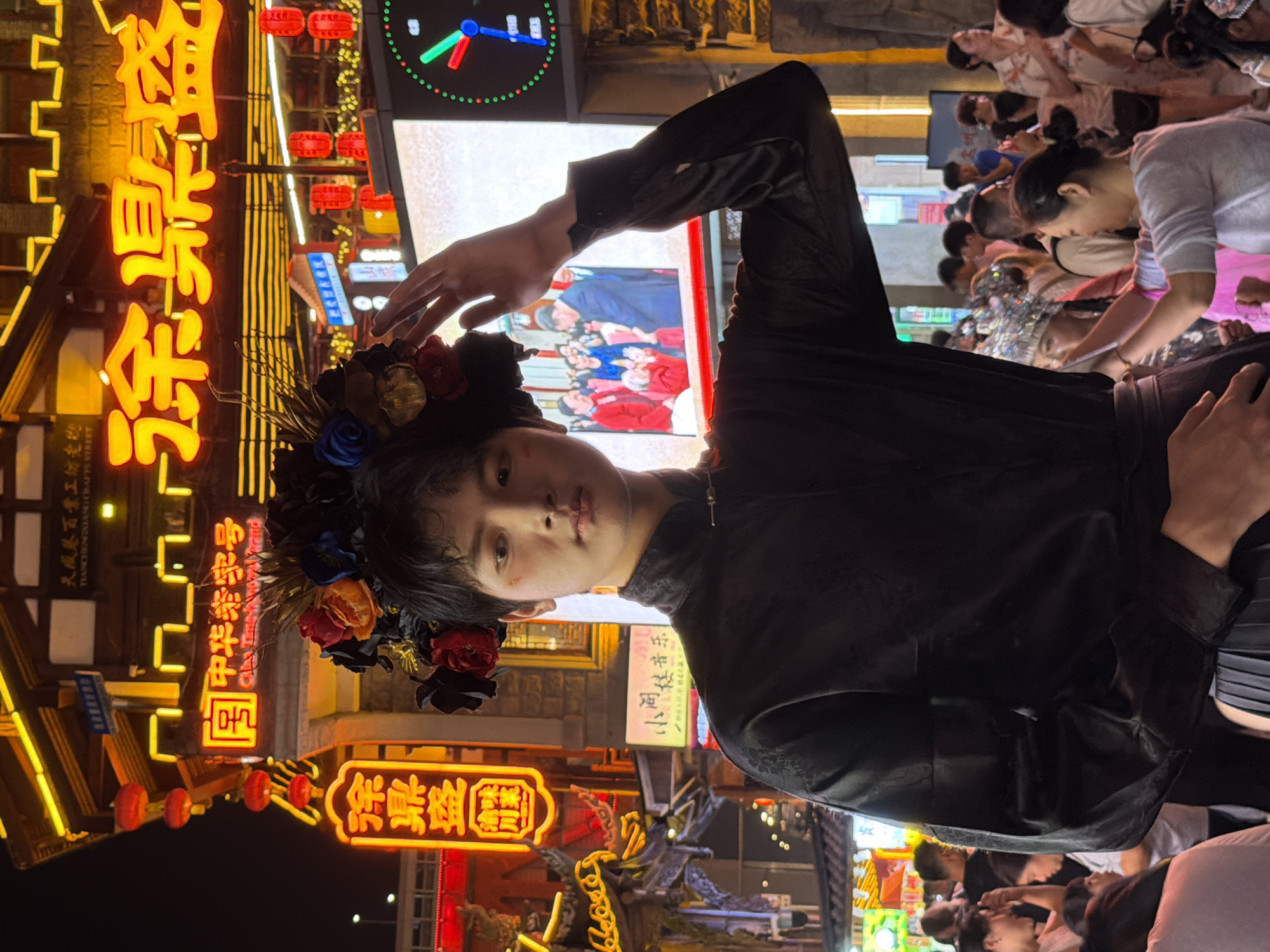 This is a photograph taken at night, likely during a festival or a busy evening in a vibrant district of Yuzhong, China, known for its crafts and traditional businesses. The image features a person in the foreground, possibly a performer or model, dressed in a dark, ornate outfit and adorned with a floral headdress. Their face is partially visible, and their hand is raised, creating a dramatic pose.
Behind the central figure, the scene is illuminated by a profusion of neon signs in Chinese characters, predominantly in yellow and red, displaying advertisements and shop names like "天成感百景工坊老物" and "中华老字号" (a designation for time-honored brands). Red lanterns are also visible, adding to the festive atmosphere. In the background, a crowd of people can be seen, some looking at displays or engaged in activities, suggesting a lively marketplace or street event. A digital clock face is also visible at the top of the frame, indicating the passage of time. The overall impression is one of a bustling, culturally rich urban environment at night, filled with light, color, and activity.